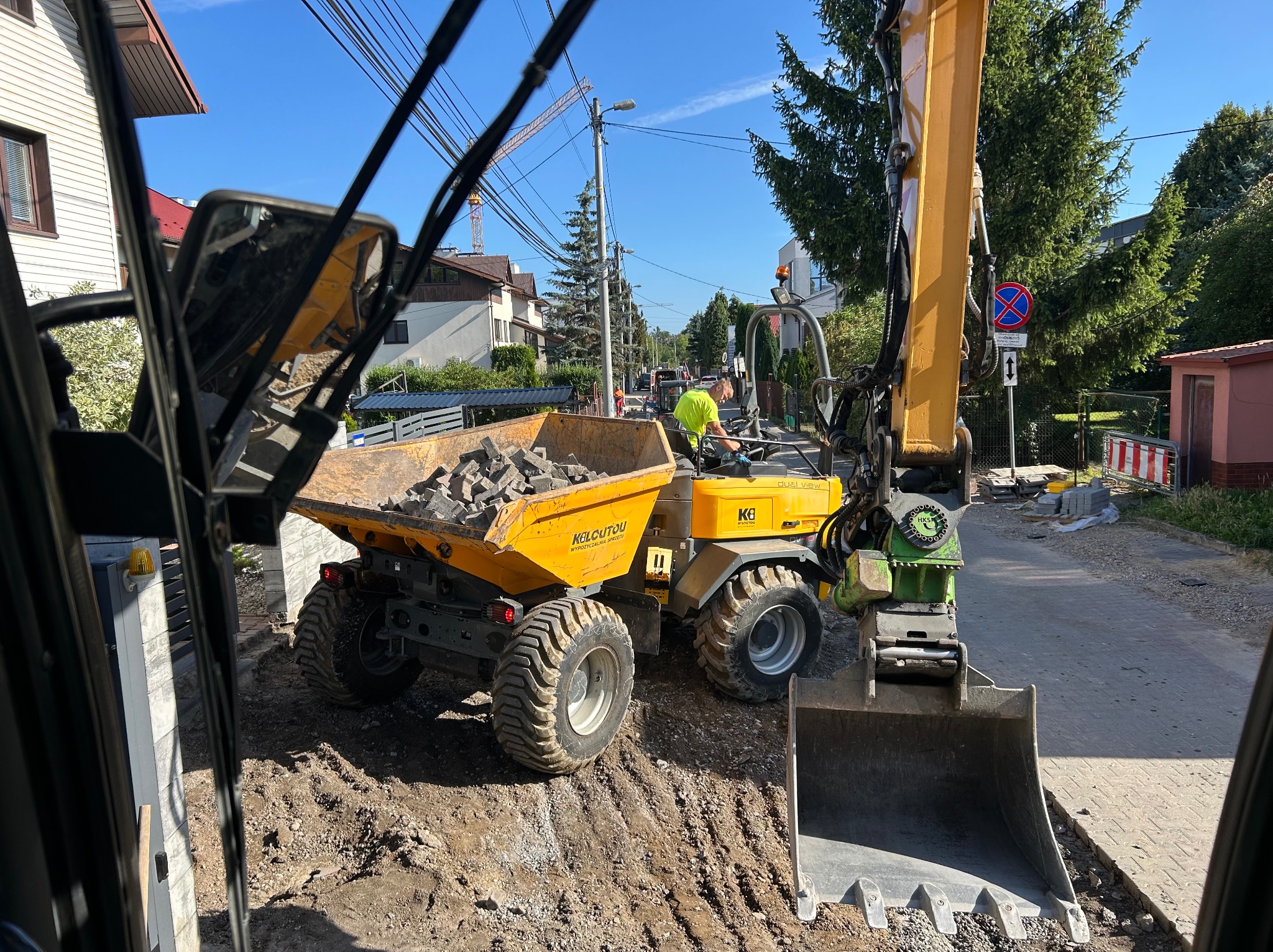 Żółta wywrotka z kostką brukową i koparka na ulicy w trakcie prac. Widok z kabiny maszyny budowlanej.
