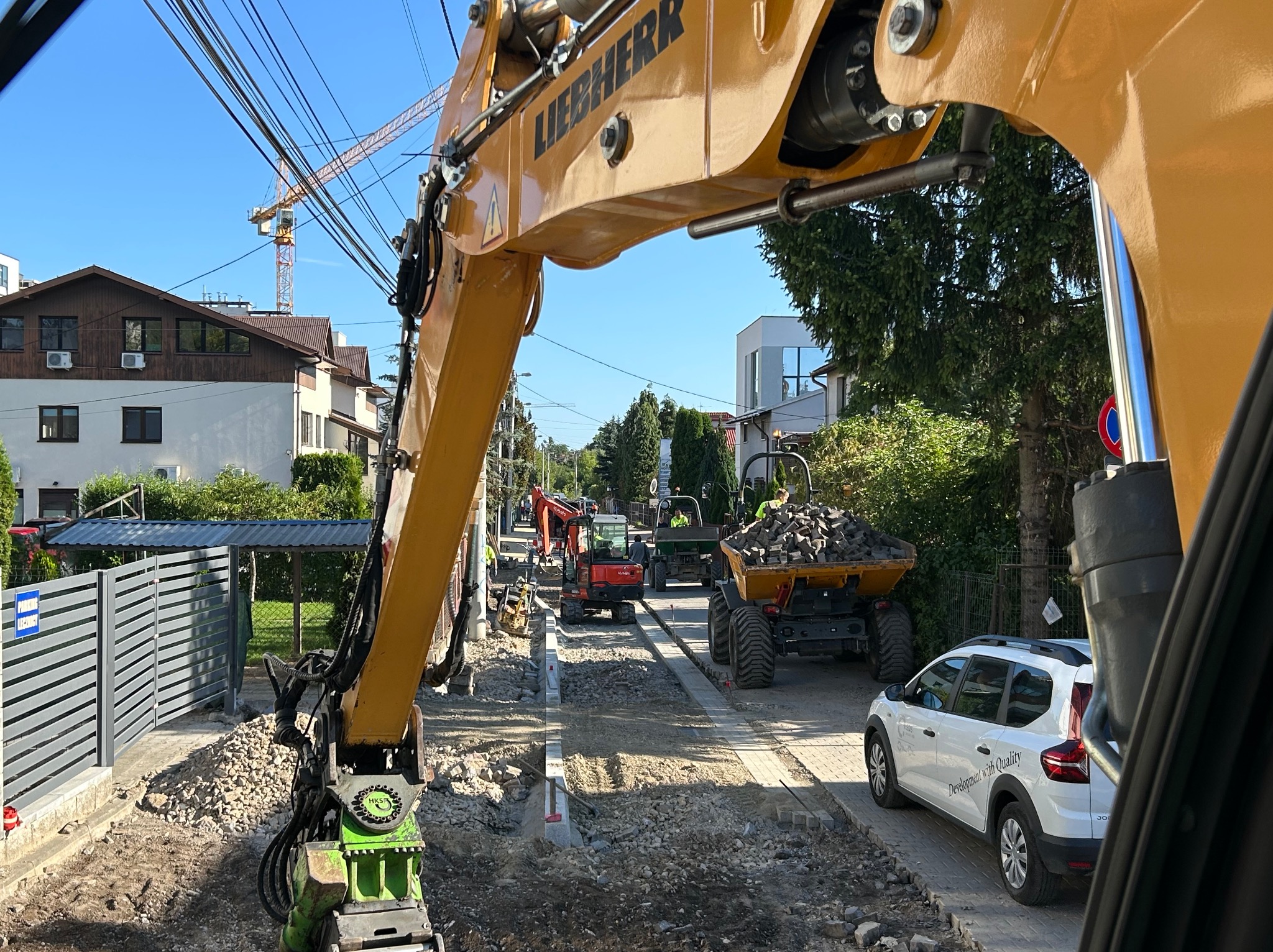 Układanie chodnika z kostki w Wola kalinowska: widok z kabiny maszyny budowlanej na postęp prac. W tle inne maszyny i budynki mieszkalne.