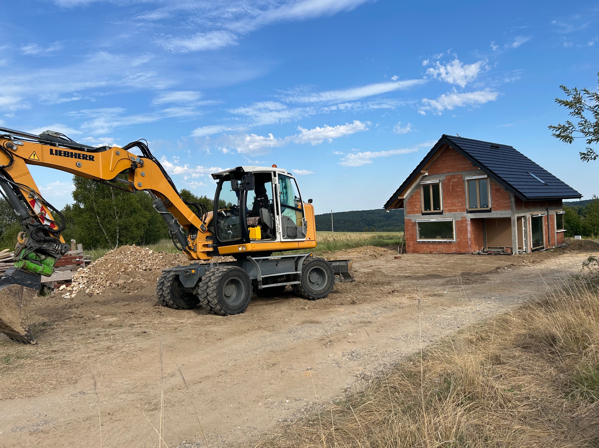 Żółta koparka Liebherr na placu budowy, w tle budynek w trakcie budowy z ciemnym dachem i licznymi oknami. Niebo błękitne z chmurami.