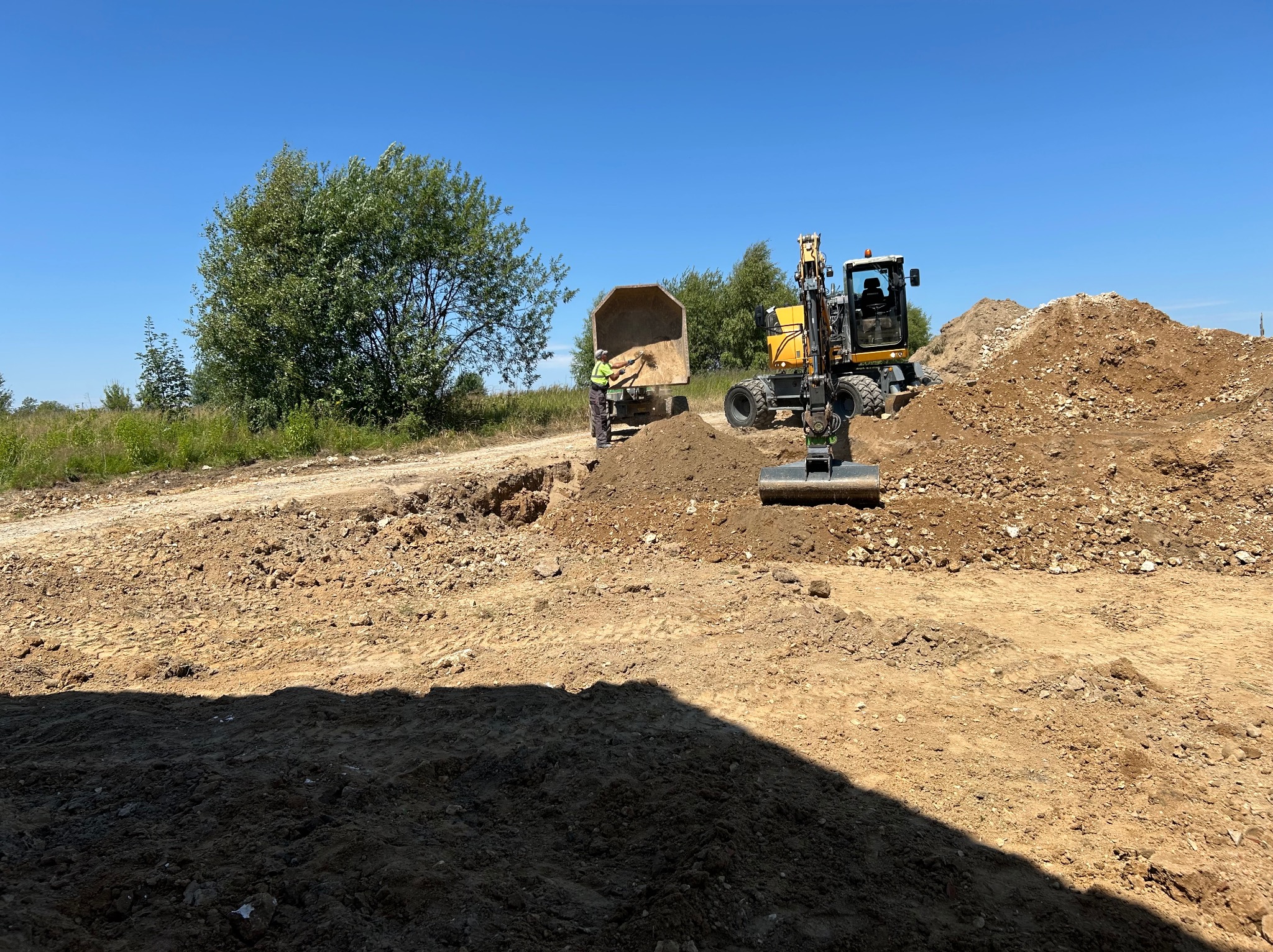 Żółta koparka kołowa i wywrotka przy pryzmach ziemi na placu budowy w słoneczny dzień. Operator przy ciężarówce, drzewo w tle. Prace ziemne w toku.