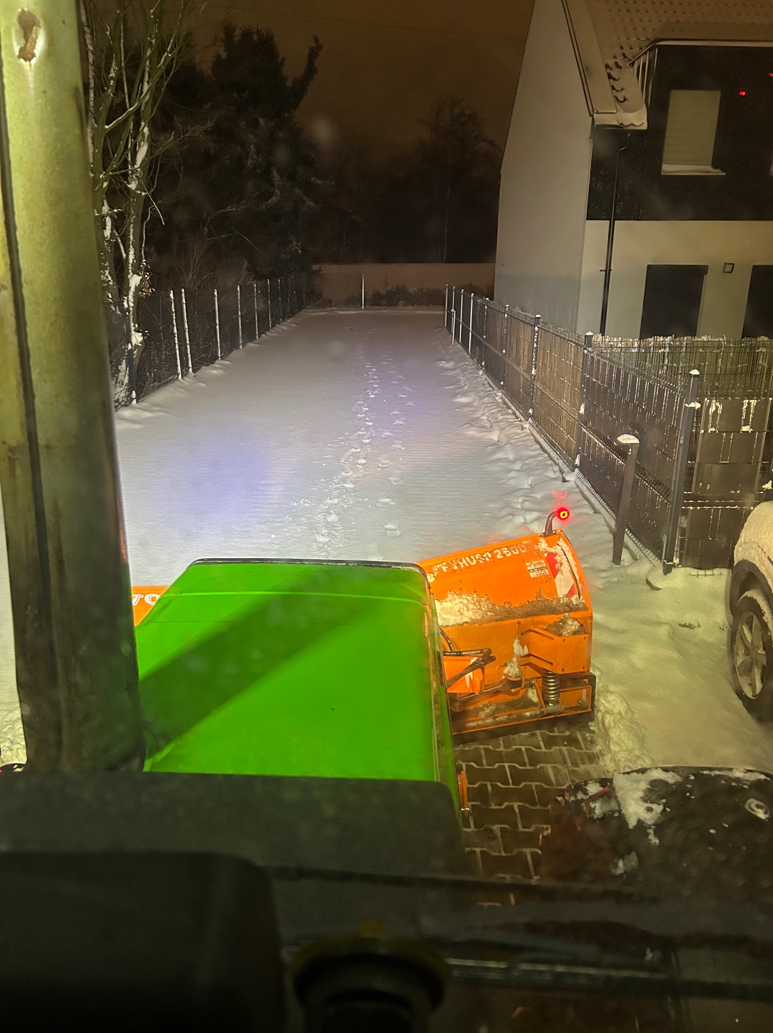 Widok z kabiny traktora z pługiem odśnieżającym, na zaśnieżoną drogę prowadzącą wzdłuż ogrodzenia posesji, nocą. Ślady na śniegu.