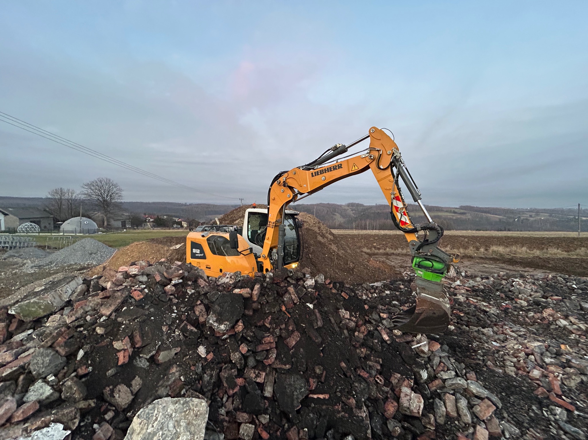 Żółta koparka Liebherr 912 compact na stercie gruzu ceglanego, w tle pole i zabudowania, pochmurne niebo. Specyficzny uchwyt koparki.