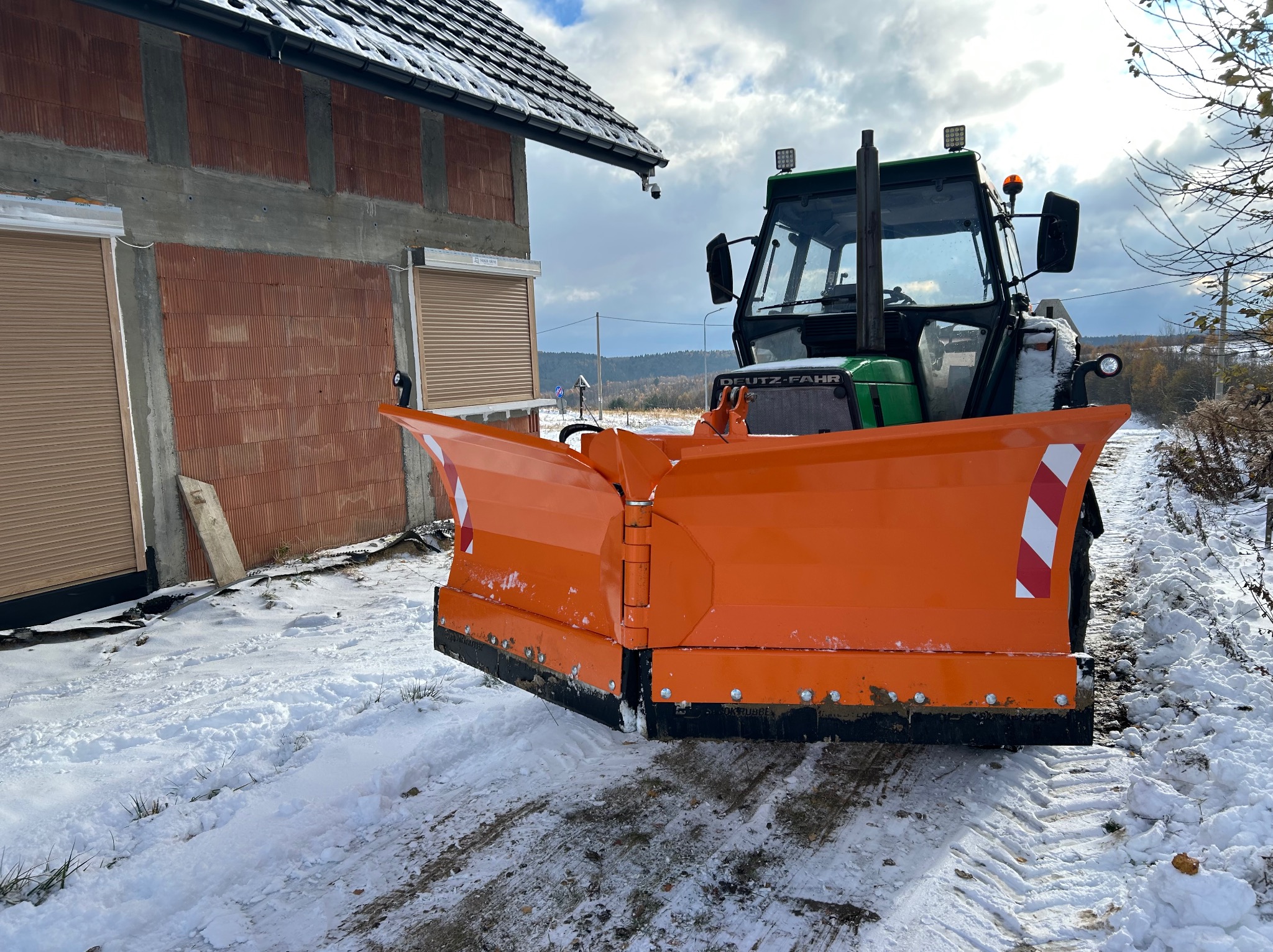 Zielony traktor z pomarańczowym pługiem odśnieżnym stoi na zaśnieżonej drodze, obok budynku w trakcie budowy. Widoczne ślady opon na śniegu.