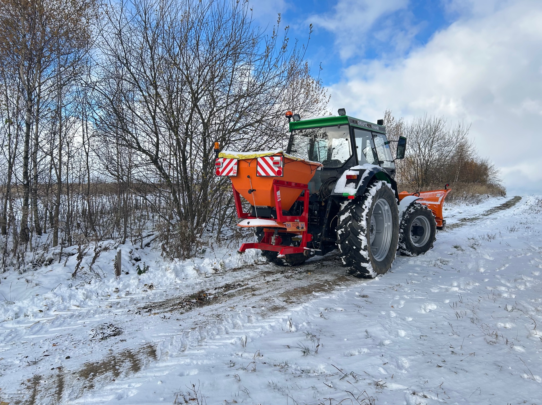 Traktor z pomarańczowym rozsiewaczem na zaśnieżonej drodze, widok z tyłu. Zimowy krajobraz z drzewami bez liści i błękitnym niebem w tle.