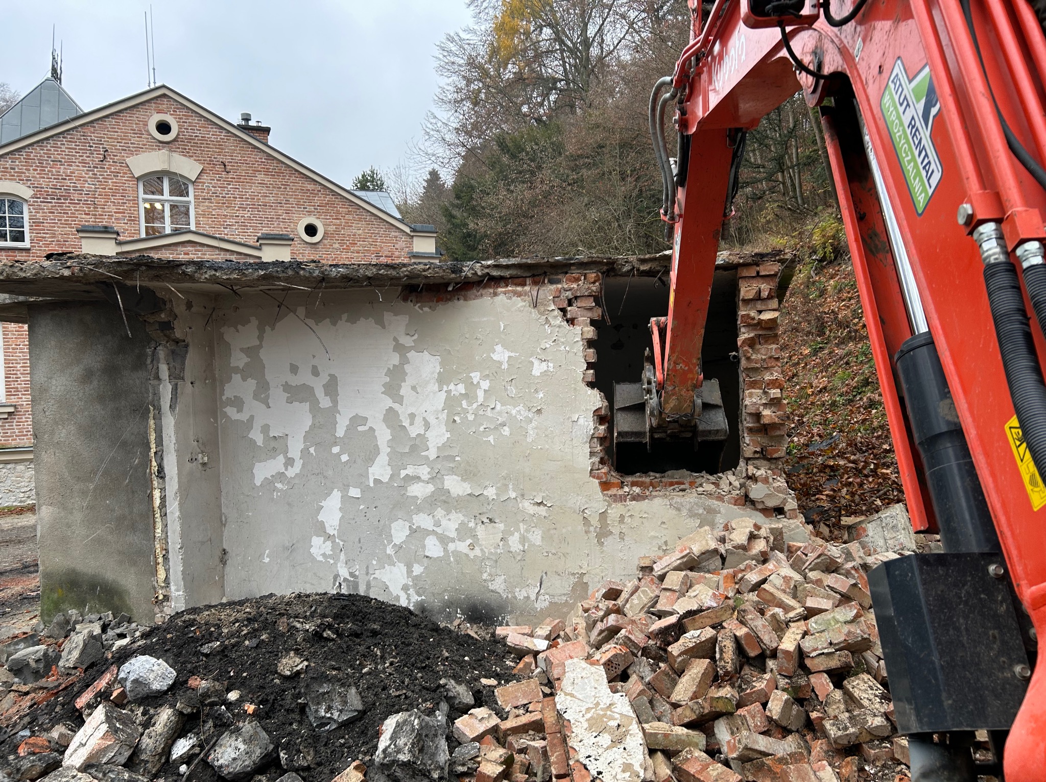 Częściowo rozebrany budynek z cegły i betonu, z widocznym ramieniem koparki. Gruz i ziemia u podstawy, budynek w tle. Etap prac rozbiórkowych w dzień.
