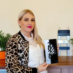 Kobieta w bluzce w panterkę trzyma nagrodę 'Orły Nieruchomości 2020' na tle biurka z dyplomem i roślinami w pomarańczowych doniczkach.