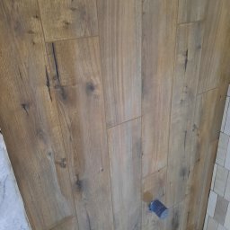 BUILD-MAX - Ściana wykończona panelami drewnopodobnymi z widocznym fragmentem instalacji wodnej i fragmentem ściany wyłożonej małymi płytkami ceramicznymi.