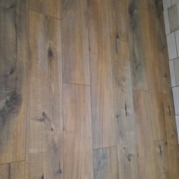 BUILD-MAX - Ułożona pod kątem brązowo-szara podłoga z paneli drewnopodobnych, widoczne sęki i usłojenie, przy krawędzi ściany wykończonej białymi płytkami.