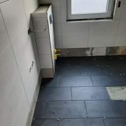 BUILD-MAX - Układanie ciemnych płytek podłogowych w łazience, widoczne krzyżyki dystansowe, białe płytki ścienne i okno dachowe w trakcie remontu.