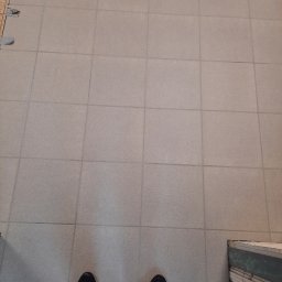 BUILD-MAX - Jasnoszare płytki podłogowe ułożone w kwadratową siatkę, widoczne stopy w czarnych butach sportowych i fragment schodów z zabezpieczeniem krawędzi.