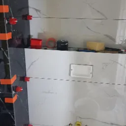 Układanie płytek imitujących marmur w łazience, z widocznymi narzędziami glazurniczymi i elementami instalacji. Częściowo ułożone płytki w kolorze czarnym.