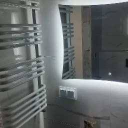 Nowoczesna łazienka z podświetlanym lustrem, grzejnikiem drabinkowym i umywalką nablatową z baterią w kolorze złotym. Płytki imitujące marmur na ścianach.