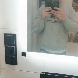 BUILD-MAX - Nowoczesna łazienka z podświetlanym lustrem, czarnymi akcentami i jasnymi płytkami. Mężczyzna robi zdjęcie w lustrze. Widoczny grzejnik drabinkowy.