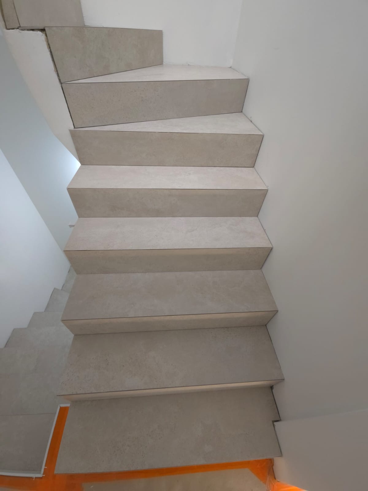 Wykończone schody wewnętrzne wyłożone jasnymi, beżowymi płytkami ceramicznymi. Widok z góry, schody prowadzą w dół, ściany białe, minimalistyczne wykończenie.