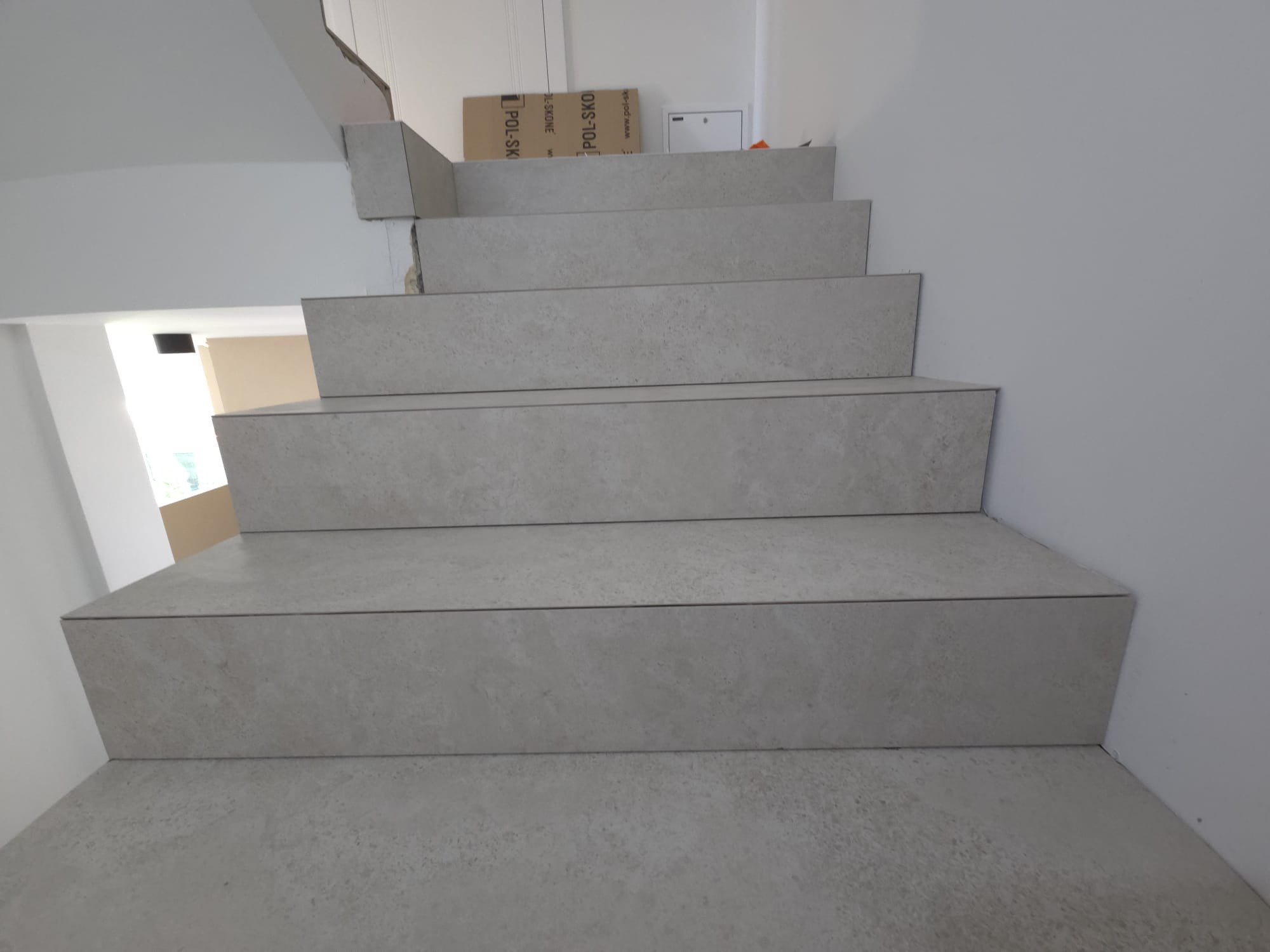 Schody wyłożone szarymi płytkami imitującymi kamień, widok z dołu. Minimalistyczne wykończenie, jasne ściany, kartonowe pudła w tle.