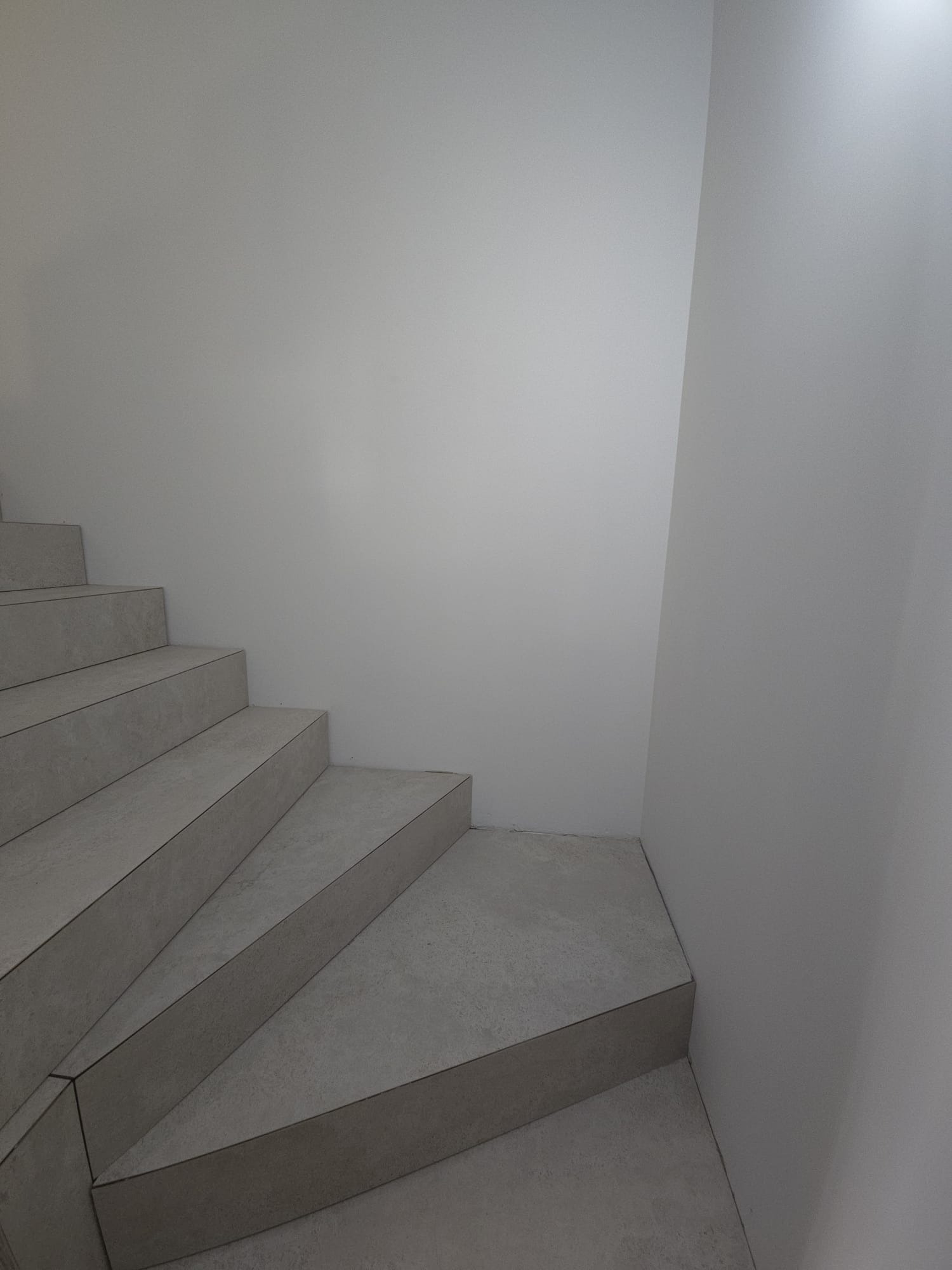 Wykończone, minimalistyczne schody wewnętrzne z szarymi płytkami, widoczne od dołu. Ściana pomalowana na biało, z delikatnym oświetleniem.