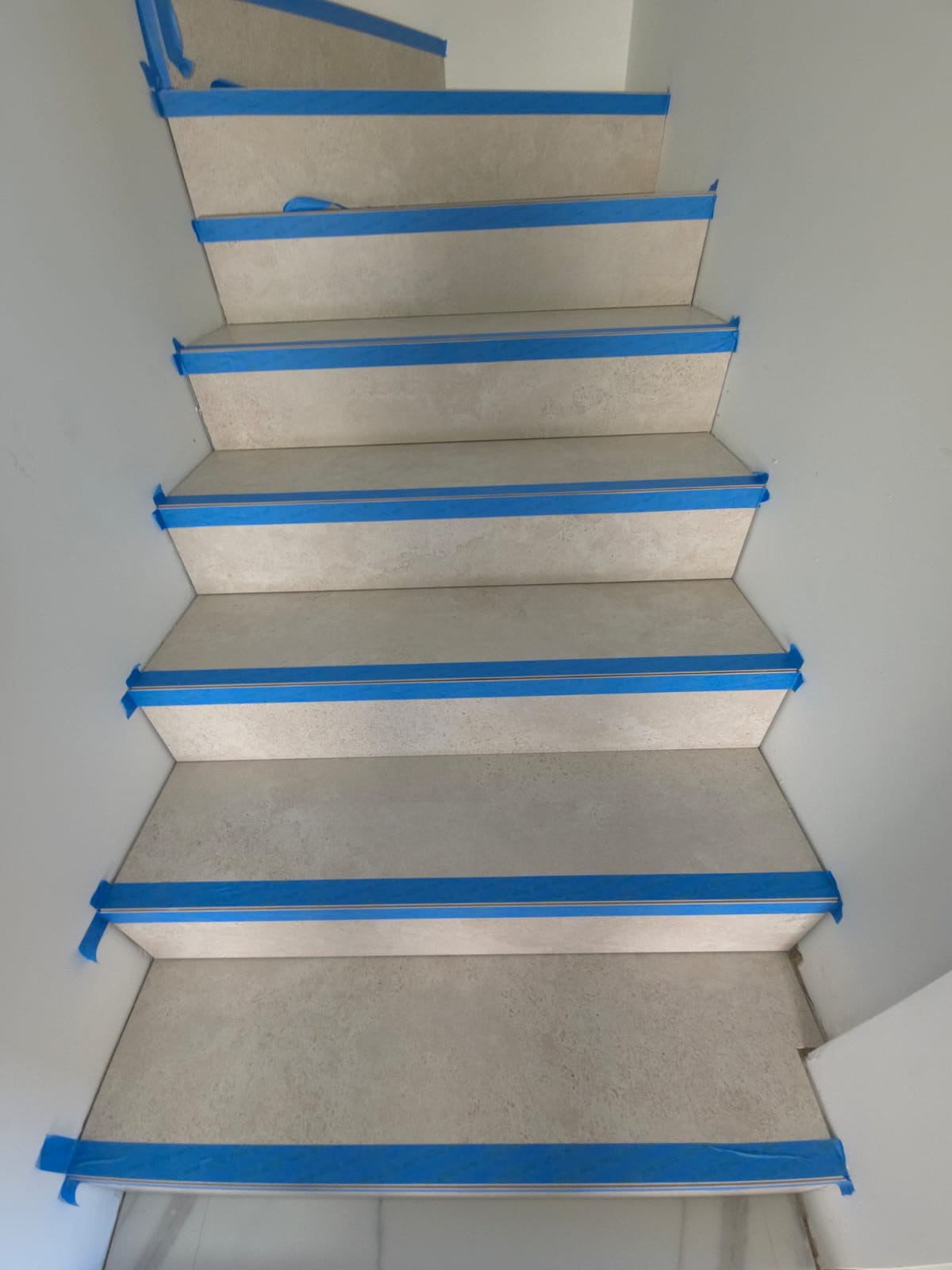 Kafelkowane schody zabezpieczone niebieską taśmą malarską, widok z góry. Jasne płytki imitujące kamień, białe ściany. Przygotowanie do fugowania lub malowania.