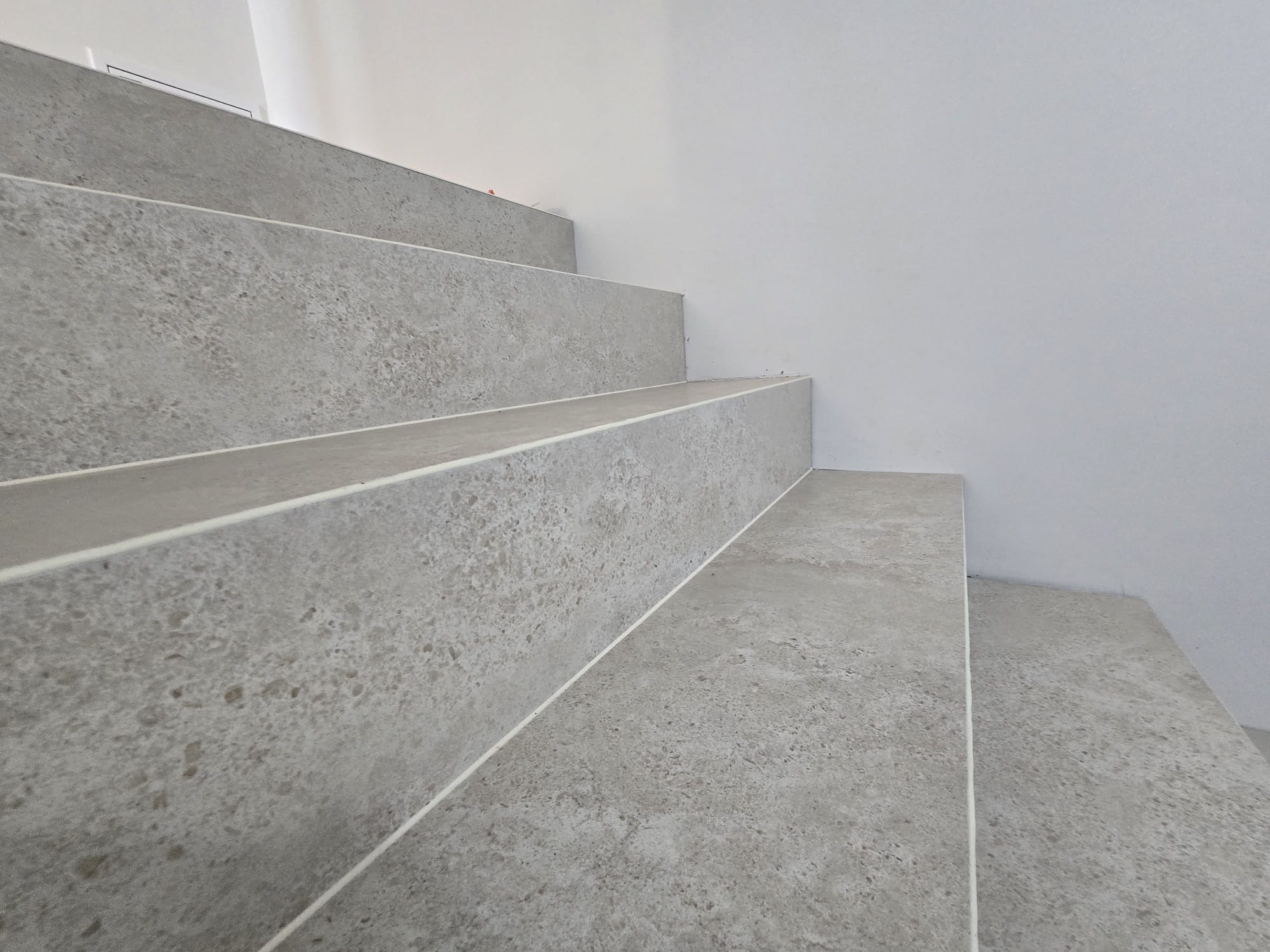 Schody wykończone szarą płytką imitującą kamień, z widocznymi fugami i białymi ścianami w tle. Minimalistyczne wykończenie.
