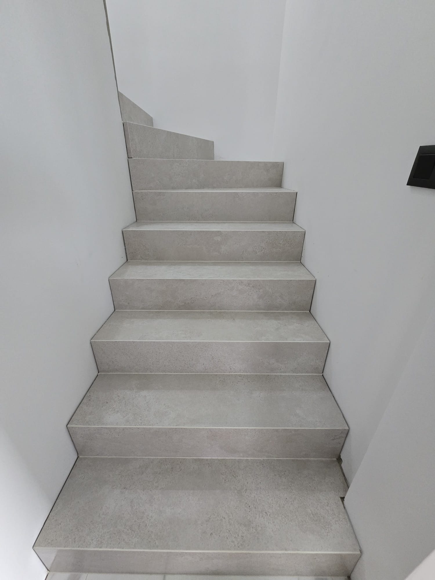 Wykończone schody wewnętrzne z szarymi płytkami, widok z dołu. Minimalistyczny design, jasne ściany, czarny włącznik na ścianie po prawej stronie.