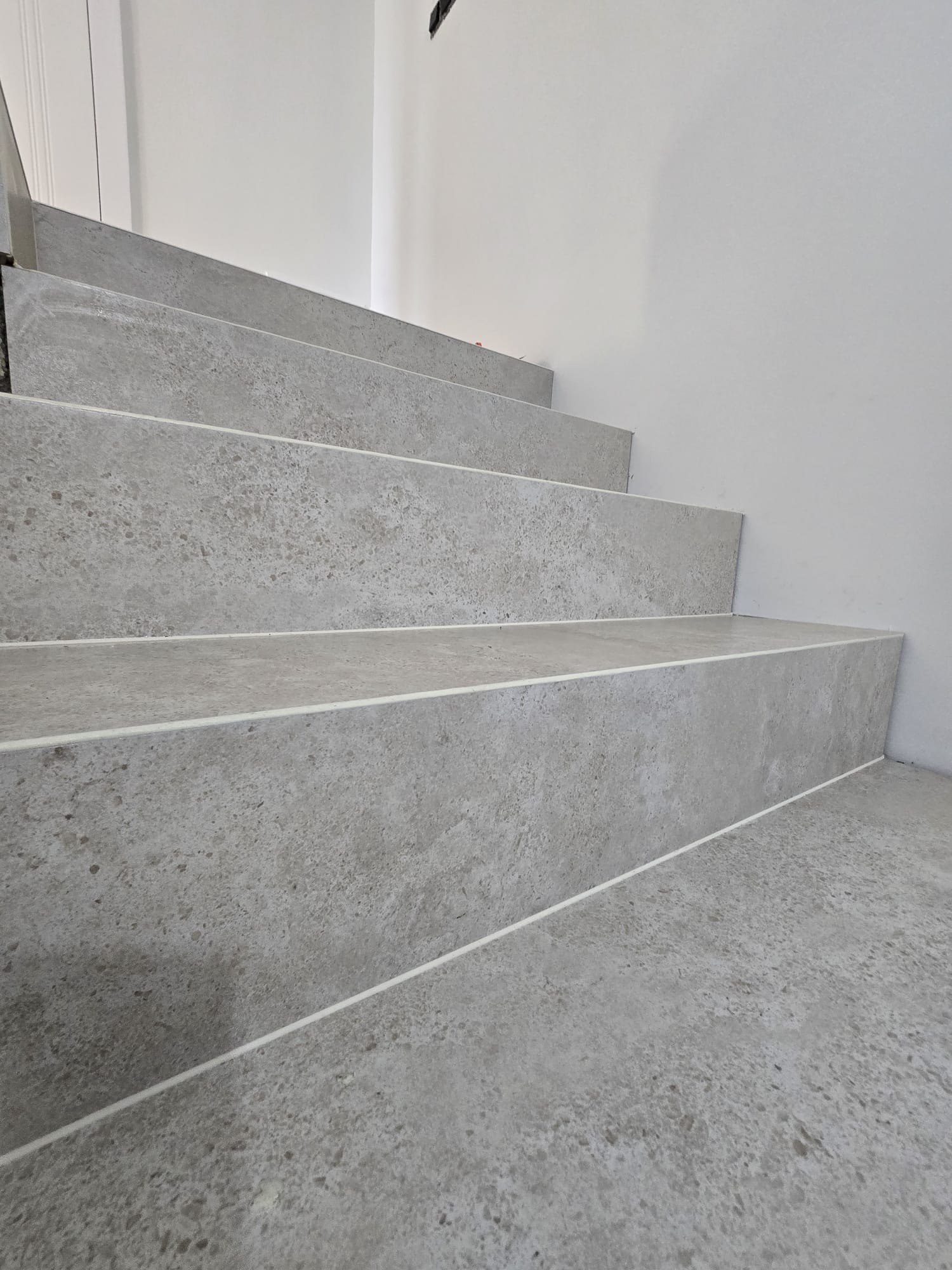 Schody wykończone szarą, kamienną płytką z jasnymi fugami, prowadzące w górę wzdłuż białej ściany. Widok z dołu, podkreślający fakturę i wykończenie.