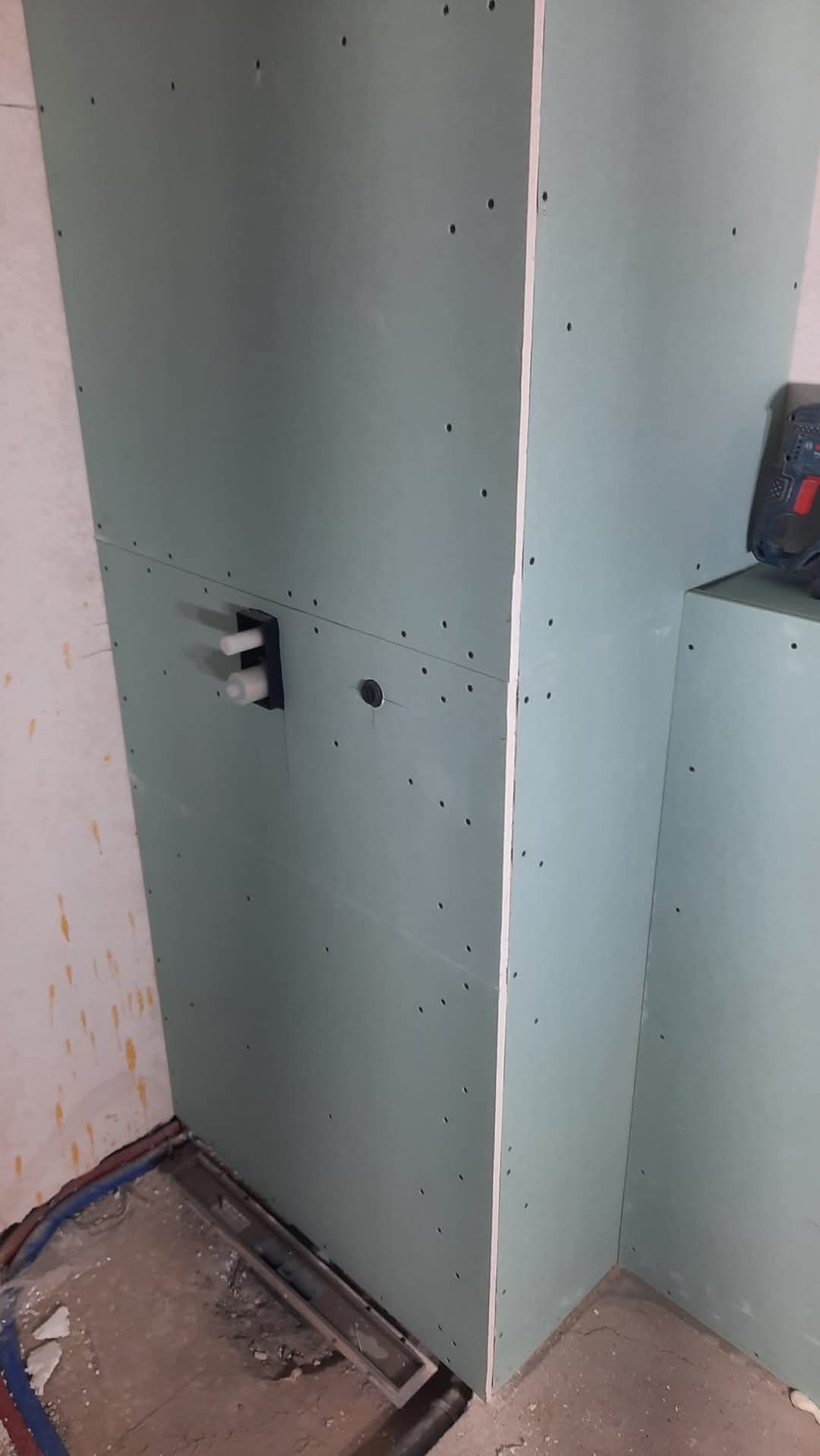 Płyta karton-gips na ścianie łazienki w trakcie montażu, widoczne przyłącza wodne i otwór pod instalację. W tle narzędzie. Prace wykończeniowe w surowym wnętrzu.