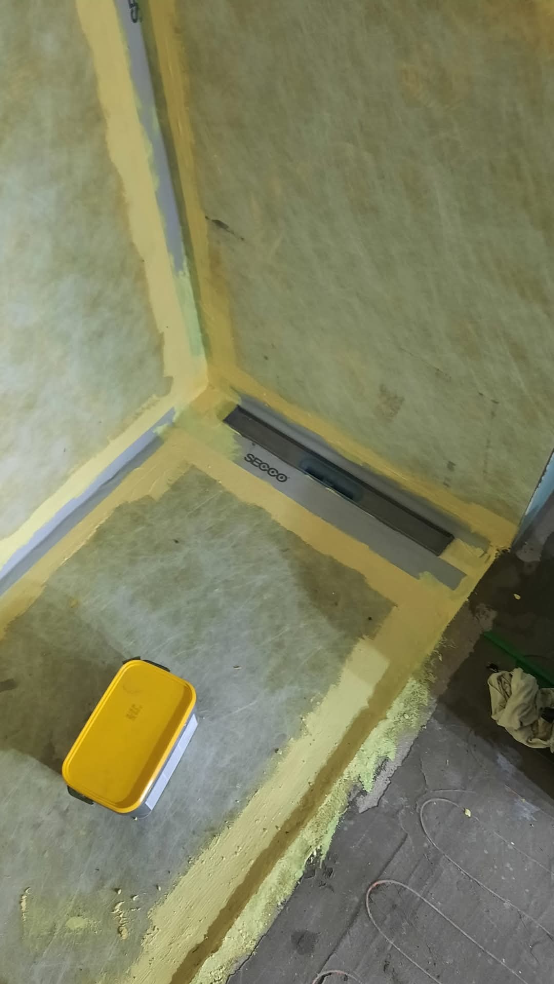 Przygotowanie do hydroizolacji w narożniku łazienki z odpływem liniowym. Żółta taśma uszczelniająca, mata izolacyjna na ścianach, ogrzewanie podłogowe.