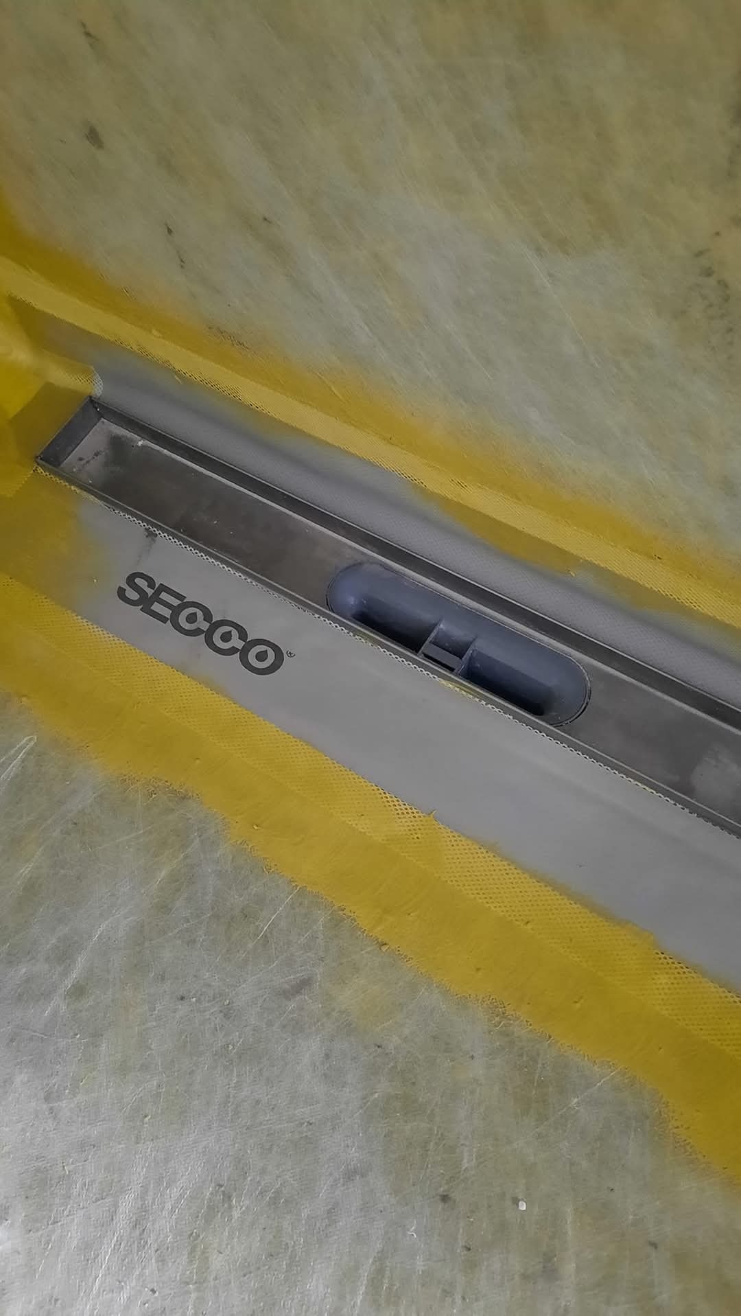 Zamontowany odpływ liniowy Secco w podłodze łazienki, zabezpieczony żółtą folią w płynie, gotowy do dalszych prac wykończeniowych. Ujęcie z góry.