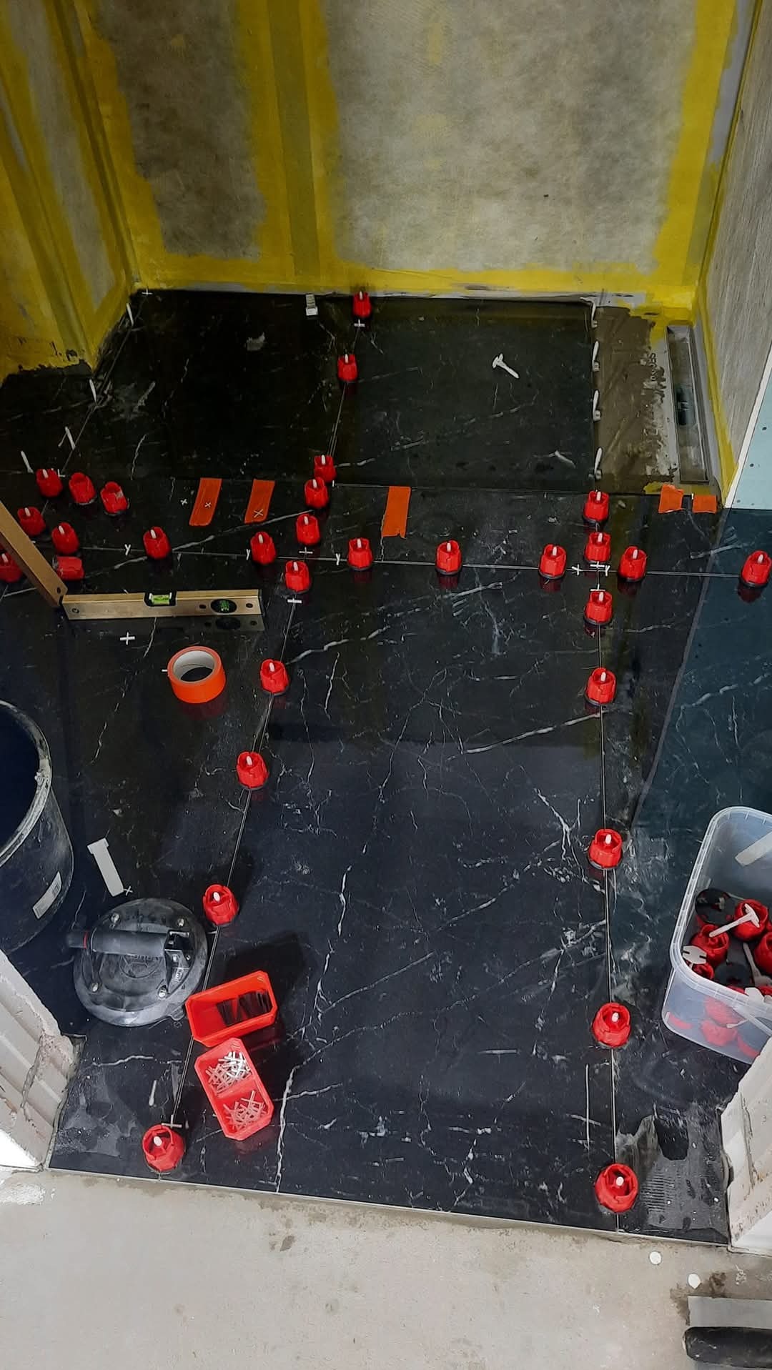 Proces układania czarnych płytek imitujących marmur w łazience, z użyciem poziomicy, klipsów i klinów do równego ułożenia. Widoczne narzędzia glazurnicze i przygotowane podłoże.