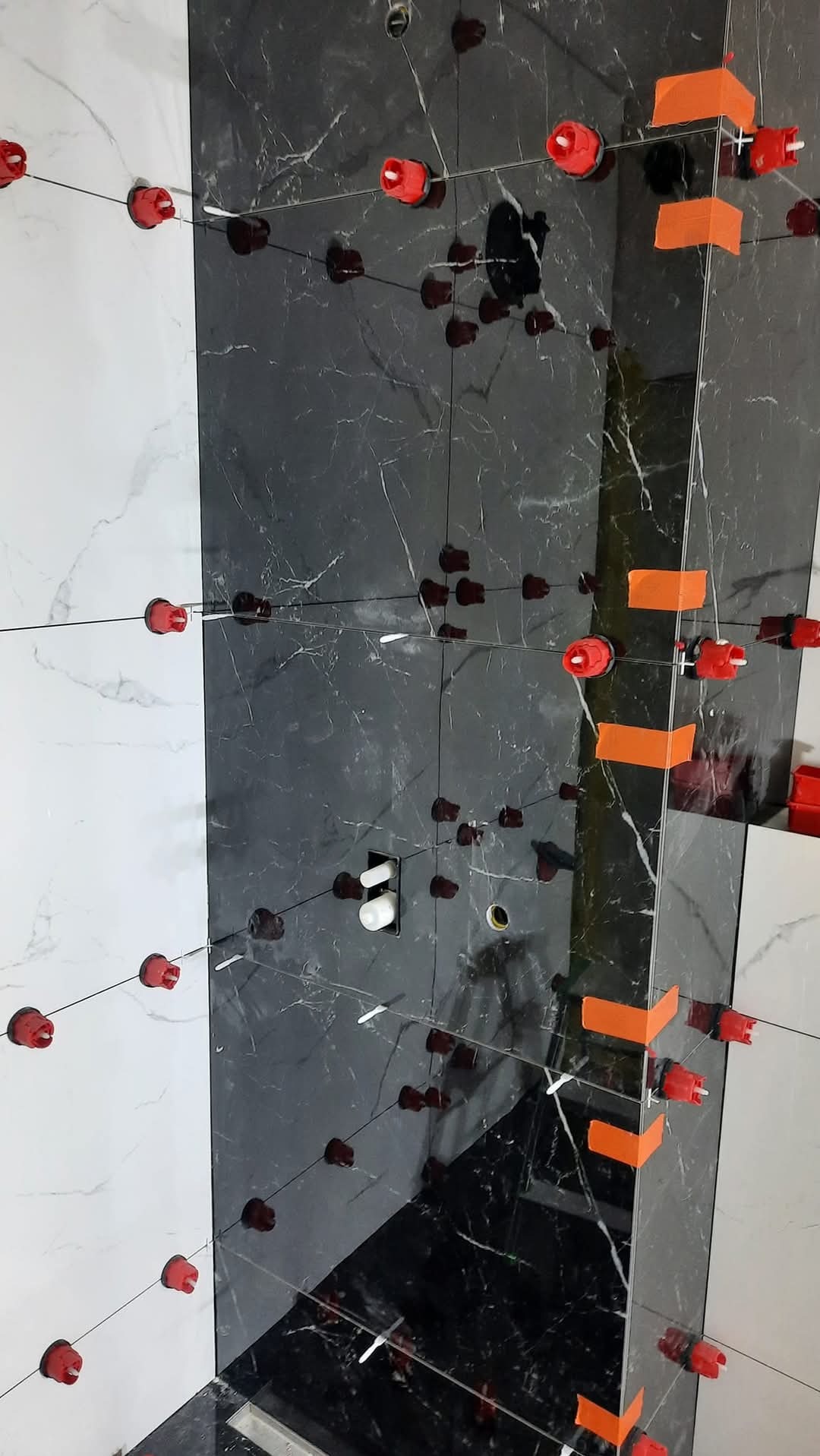 Układanie płytek w łazience z użyciem systemu poziomowania. Widoczne czarne i białe płytki imitujące marmur oraz czerwone klipsy i pomarańczowe paski dystansowe.