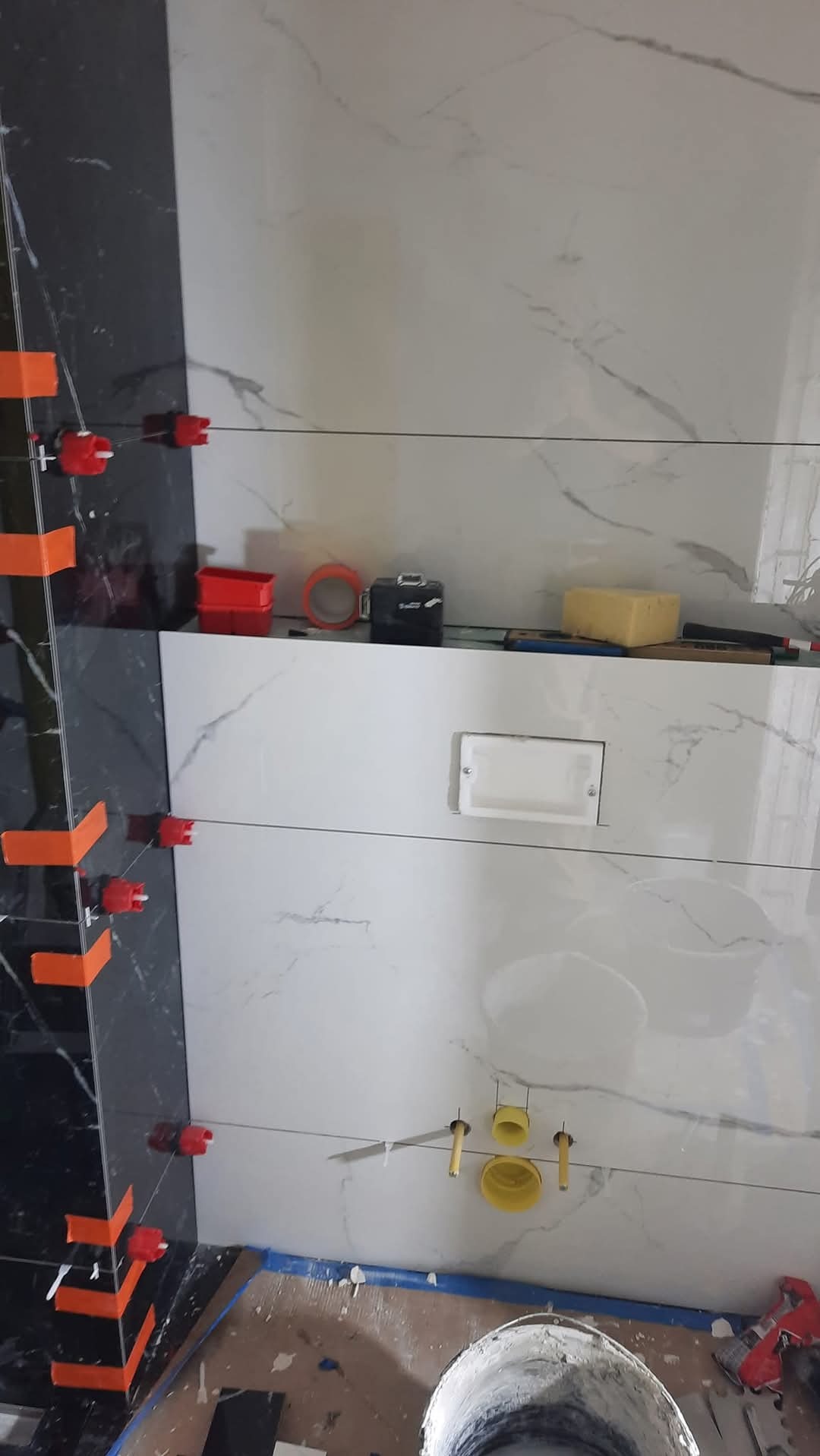 Układanie płytek imitujących marmur w łazience, z widocznymi narzędziami glazurniczymi i elementami instalacji. Częściowo ułożone płytki w kolorze czarnym.