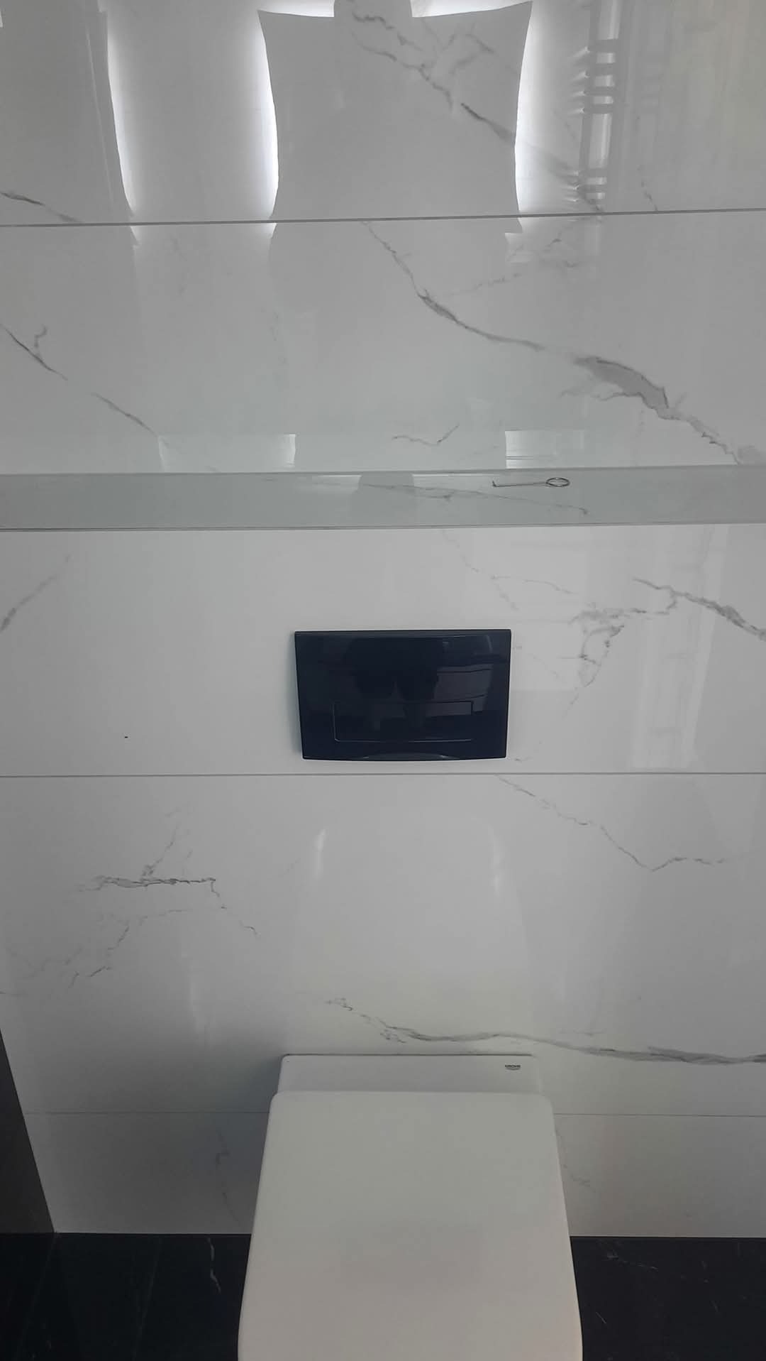 Biała toaleta podwieszana z czarnym przyciskiem spłukującym, otoczona marmurowymi płytkami w łazience. Minimalistyczny design, nowoczesne wykończenie.
