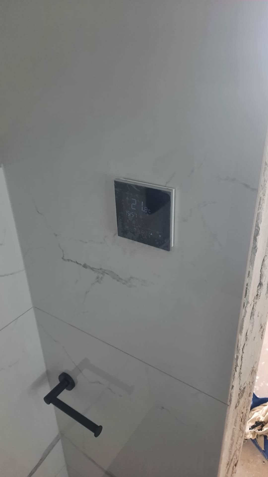 Wykończona łazienka z płytkami imitującymi marmur i nowoczesnym panelem sterowania ogrzewaniem podłogowym. Widoczny uchwyt na papier toaletowy w kolorze czarnym.