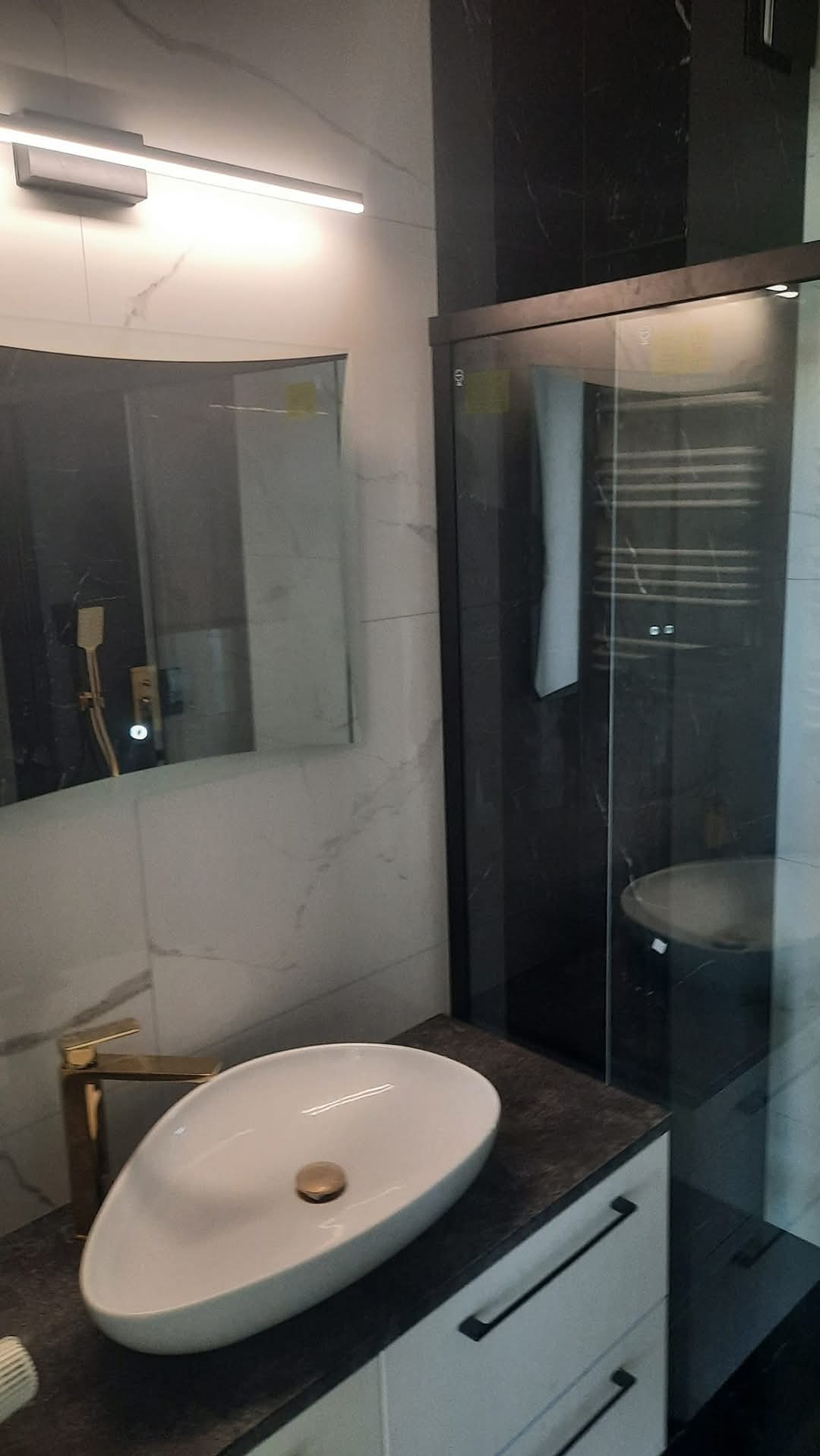 Nowoczesna łazienka z owalną białą umywalką, złotą baterią i kabiną prysznicową z czarnymi ramami na tle marmurowych płytek.