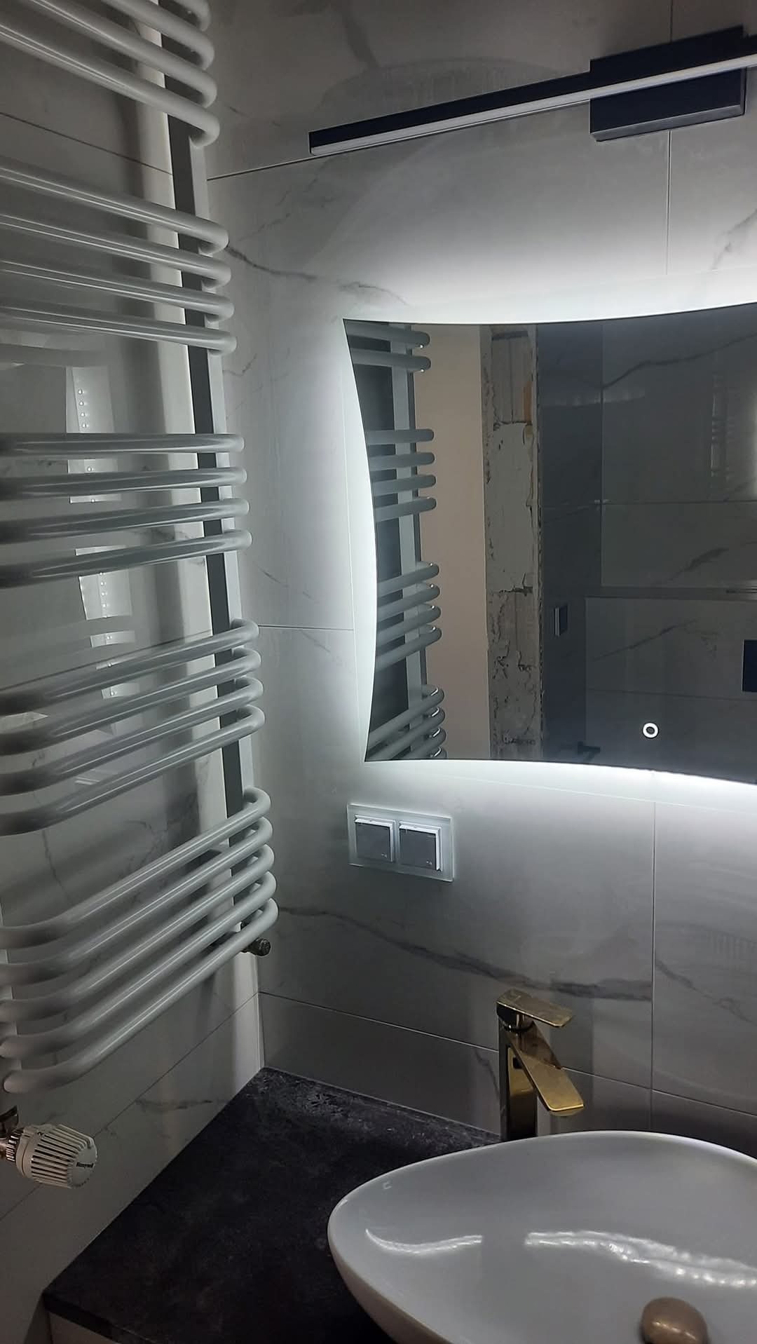 Nowoczesna łazienka z podświetlanym lustrem, grzejnikiem drabinkowym i umywalką nablatową z baterią w kolorze złotym. Płytki imitujące marmur na ścianach.