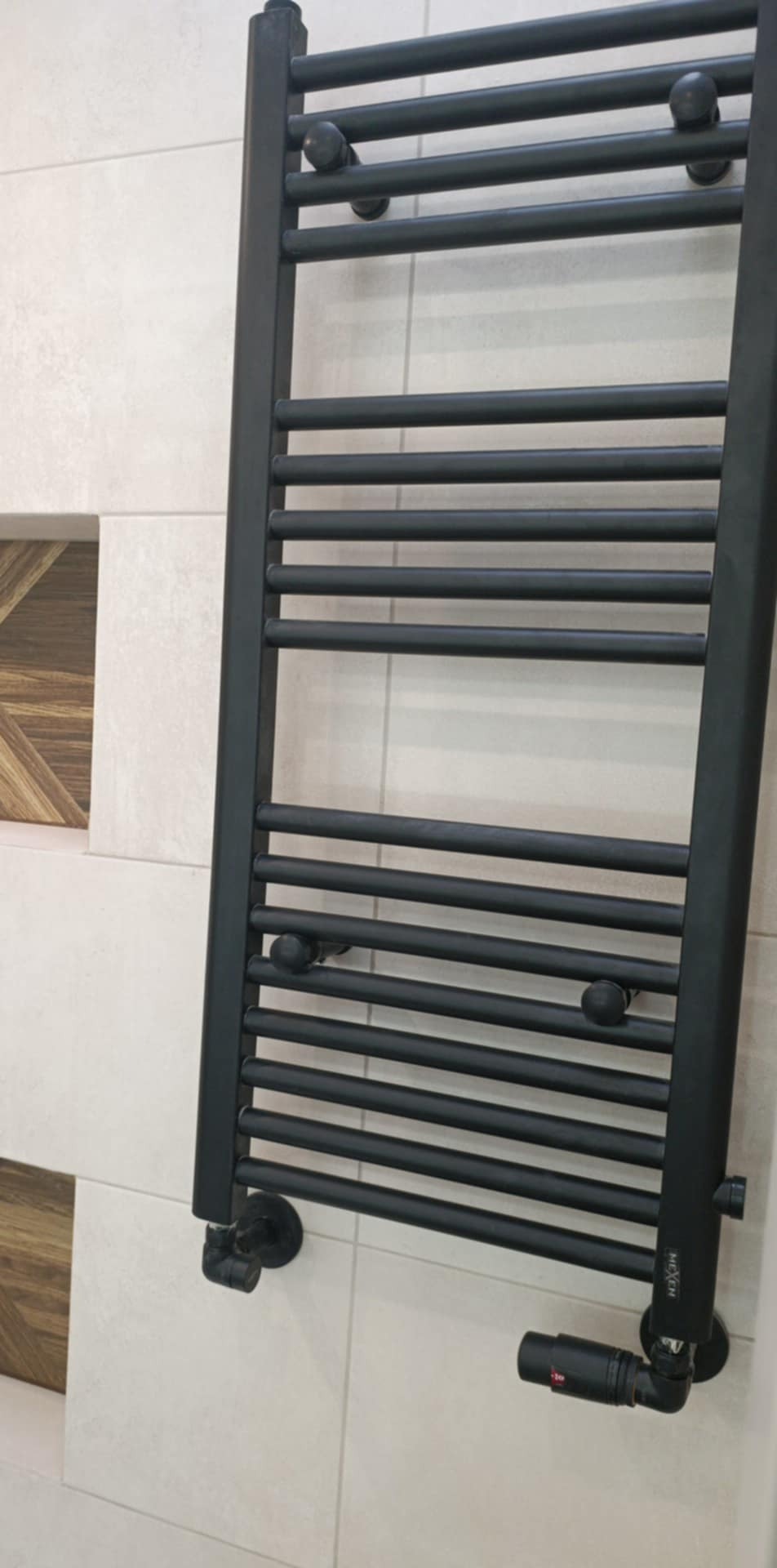 Czarny, drabinkowy grzejnik łazienkowy na tle jasnych, kwadratowych płytek z dekoracyjnym, drewnopodobnym akcentem. Widoczny zawór termostatyczny.