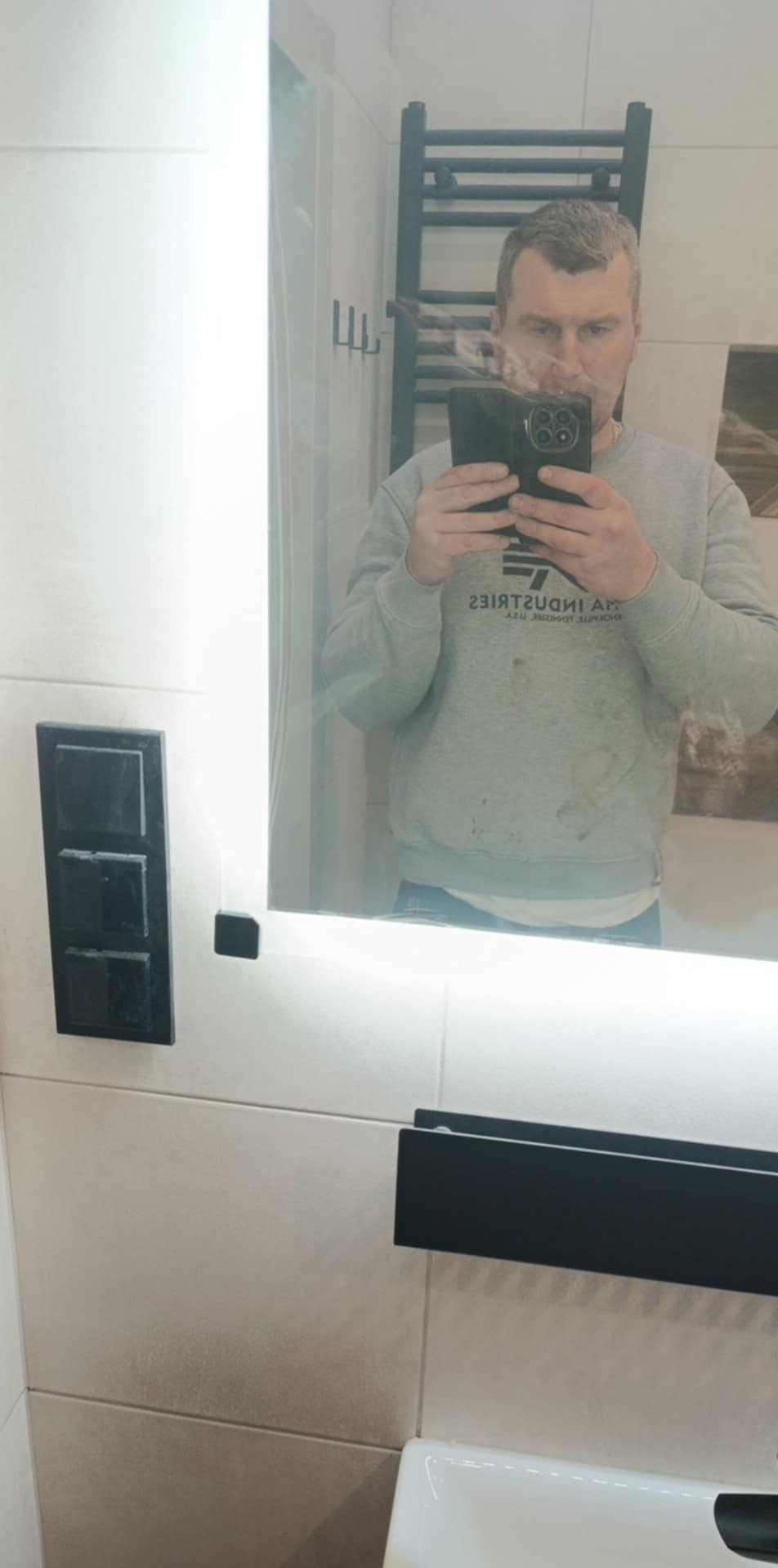 Nowoczesna łazienka z podświetlanym lustrem, czarnymi akcentami i jasnymi płytkami. Mężczyzna robi zdjęcie w lustrze. Widoczny grzejnik drabinkowy.