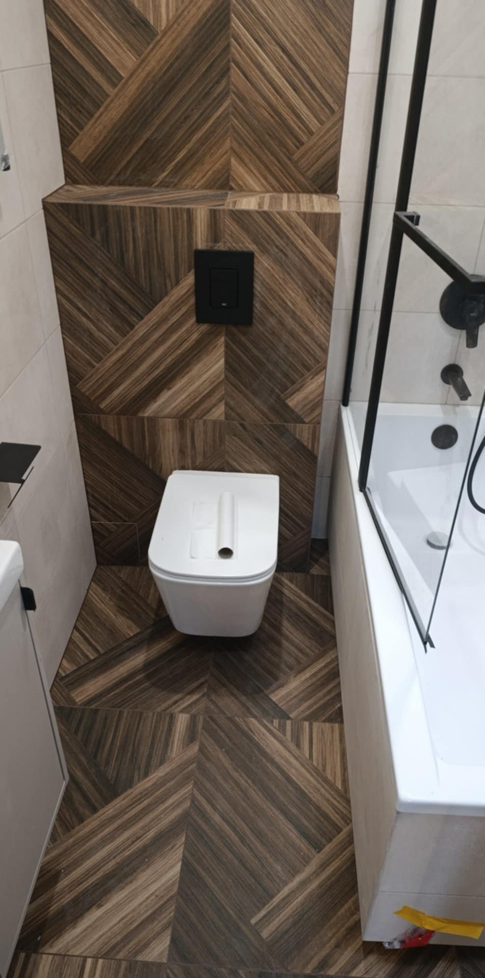 Nowoczesna łazienka z drewnopodobnymi płytkami ułożonymi w geometryczny wzór na podłodze i ścianie za toaletą. Minimalistyczna armatura w kolorze czarnym.