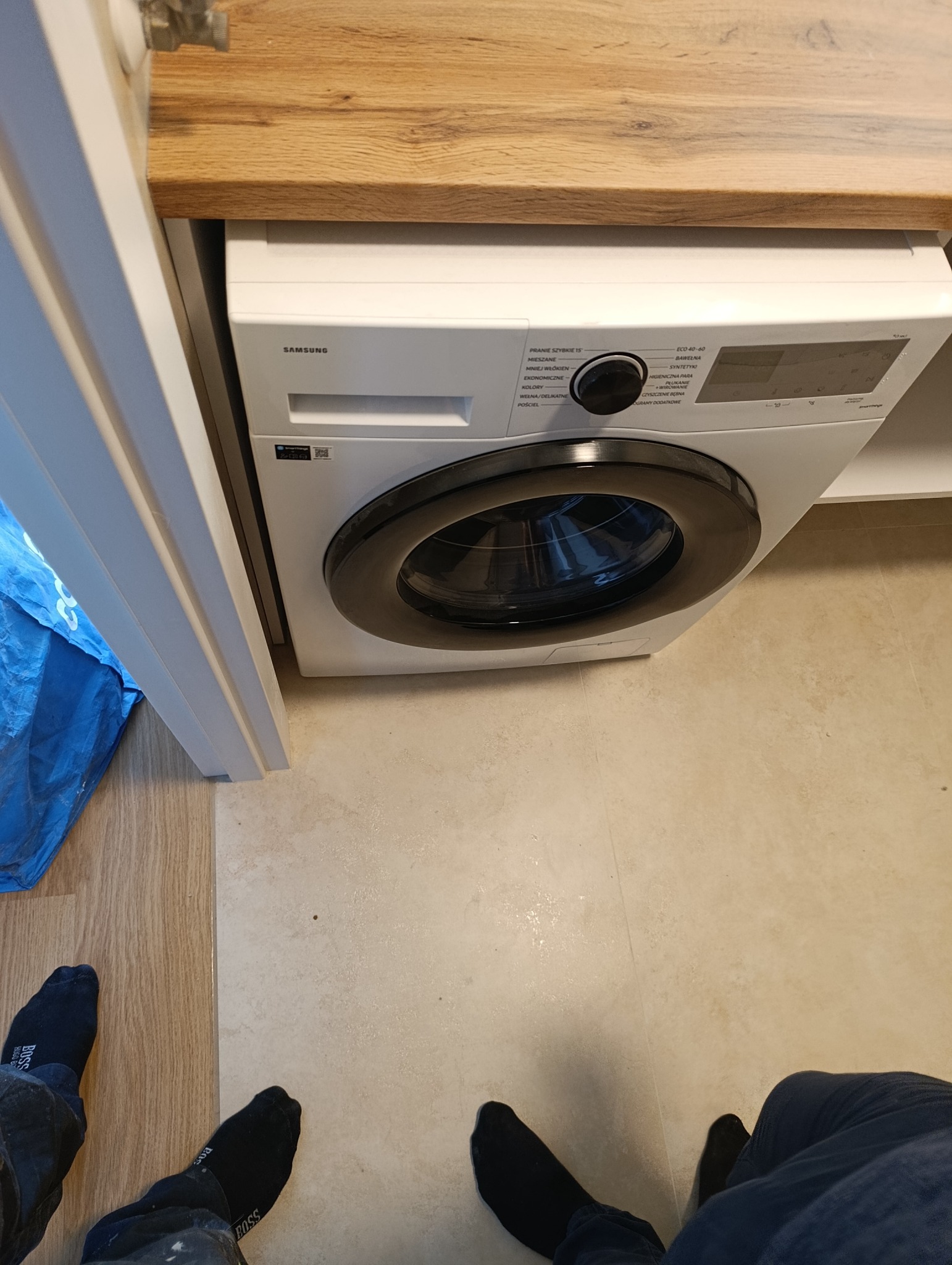 Nowoczesna pralka Samsung w zabudowie łazienkowej z drewnianym blatem. Widoczne stopy osoby robiącej zdjęcie. Jasne płytki podłogowe.