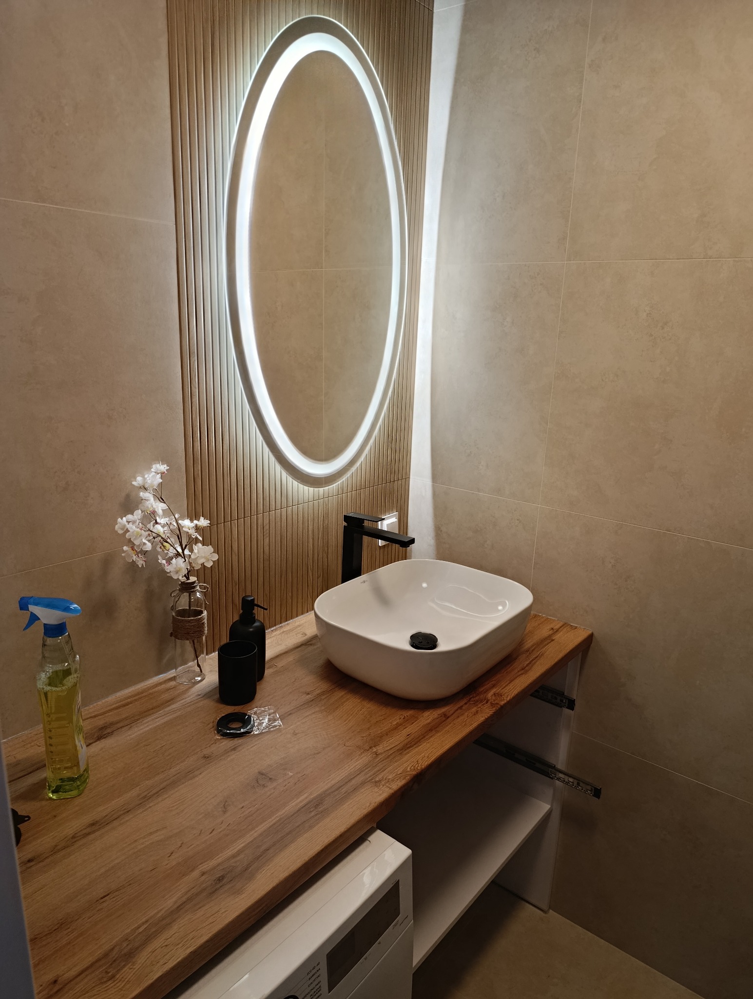 Nowoczesna łazienka z owalnym lustrem LED, drewnianym blatem i białą umywalką. Beżowe płytki na ścianach. Wazonik z kwiatami i czarne akcesoria.