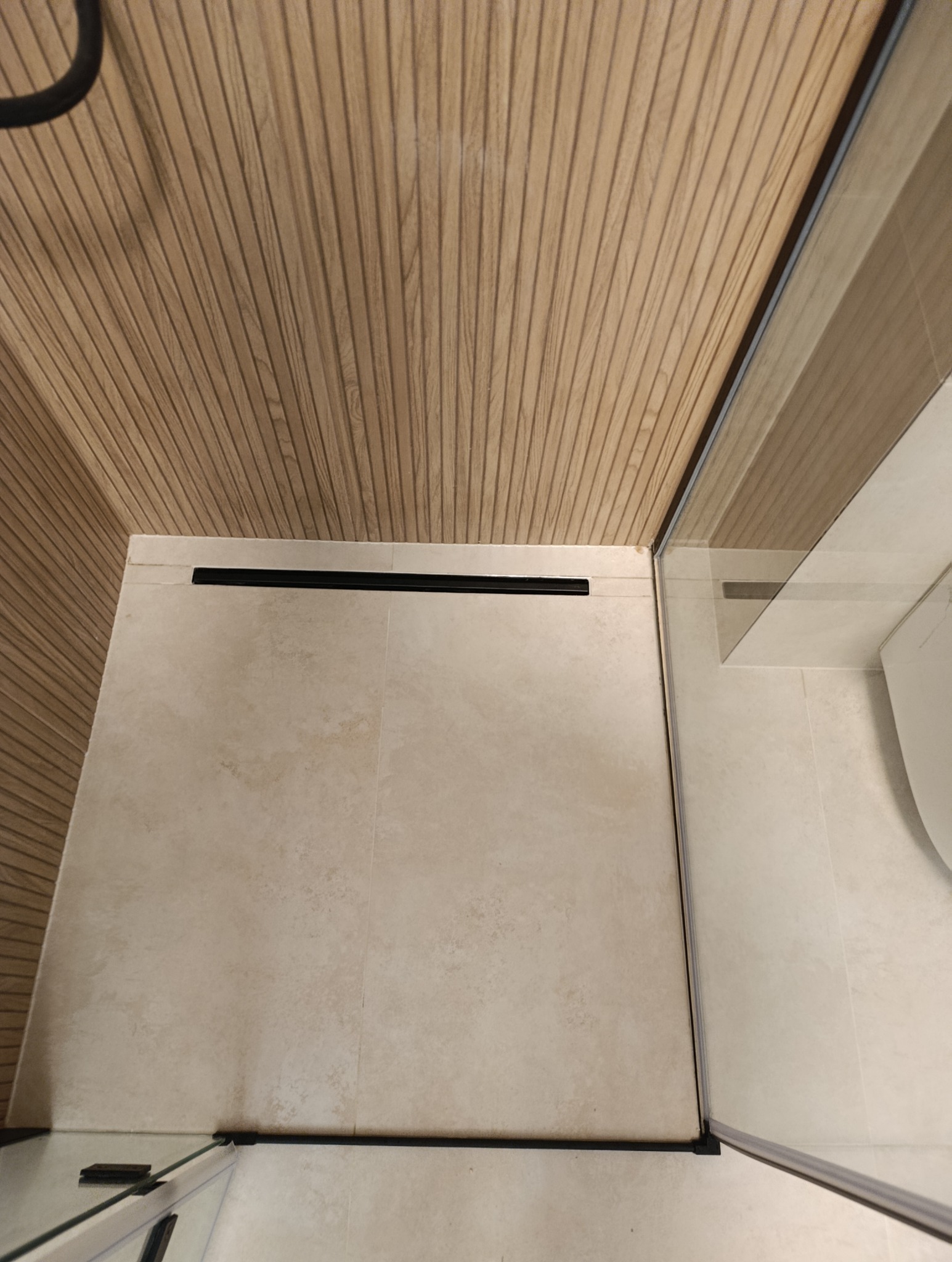 Beżowe płytki podłogowe w kabinie prysznicowej z odpływem liniowym i drewnopodobną okładziną ścienną. Widok z góry, fragment kabiny.