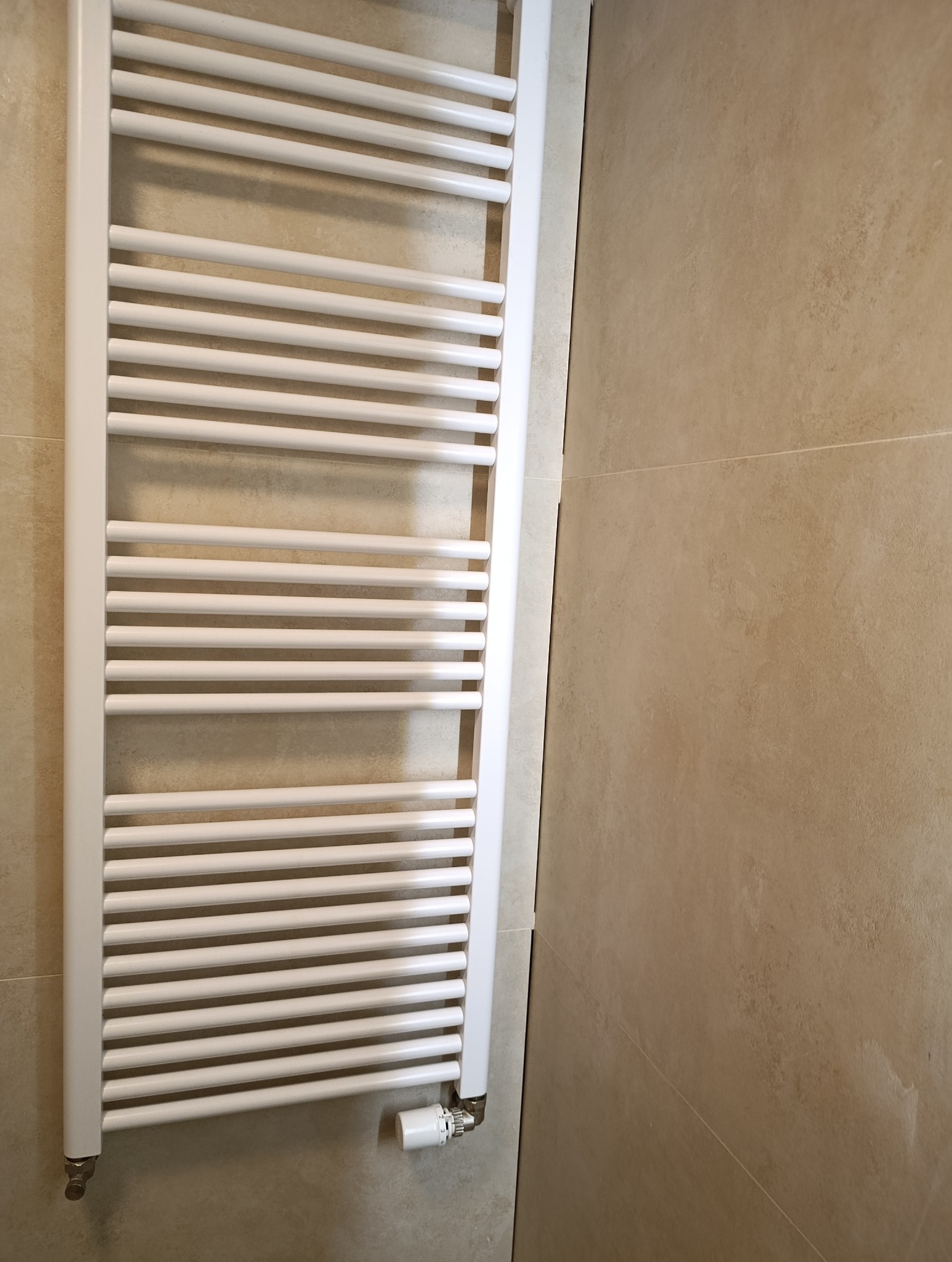 Biały, drabinkowy grzejnik łazienkowy zamontowany na beżowej, ceramicznej ścianie. Widoczny zawór termostatyczny. Ujęcie z bliska, prezentujące detale instalacji.