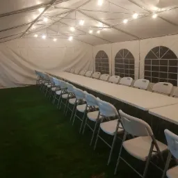 Wnętrze białego namiotu imprezowego z długim stołem i krzesłami ustawionymi na sztucznej trawie, oświetlone girlandą żarówek.