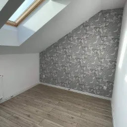 Pokój na poddaszu po remoncie: drewnopodobne panele podłogowe, ściana z szarą tapetą w kwiaty i ptaki, białe ściany, okno dachowe.