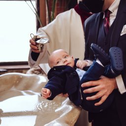 Foto Artist Fotografia Monika Jachowicz - Niemowlę w granatowym ubranku trzymane nad marmurową chrzcielnicą podczas ceremonii, ksiądz w tle polewa wodą z metalowej miseczki.