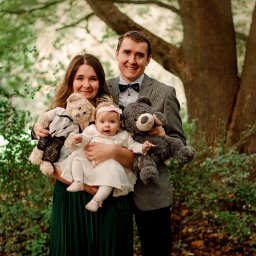 Foto Artist Fotografia Monika Jachowicz - Rodzinne zdjęcie w plenerze: rodzice trzymają dziecko i dwa pluszowe misie na tle drzew i zieleni.