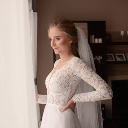 Foto Artist Fotografia Monika Jachowicz - Panna młoda w koronkowej sukni z długim rękawem i welonem, spogląda przez okno, trzymając rękę na biodrze, w tle regał z książkami i zdjęciami.