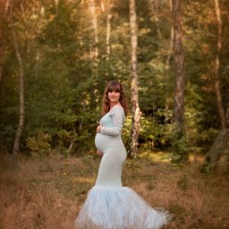Foto Artist Fotografia Monika Jachowicz - Kobieta w ciąży w długiej, błękitnej sukni z koronkowymi rękawami, pozuje w jesiennym lesie, trzymając ręce na brzuchu.