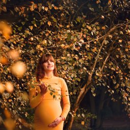 Foto Artist Fotografia Monika Jachowicz - Uśmiechnięta kobieta w dopasowanej, żółtej sukience ciążowej, trzymająca dłoń na brzuchu, pozuje pod drzewem z jesiennymi liśćmi w ciepłym, naturalnym świetle.