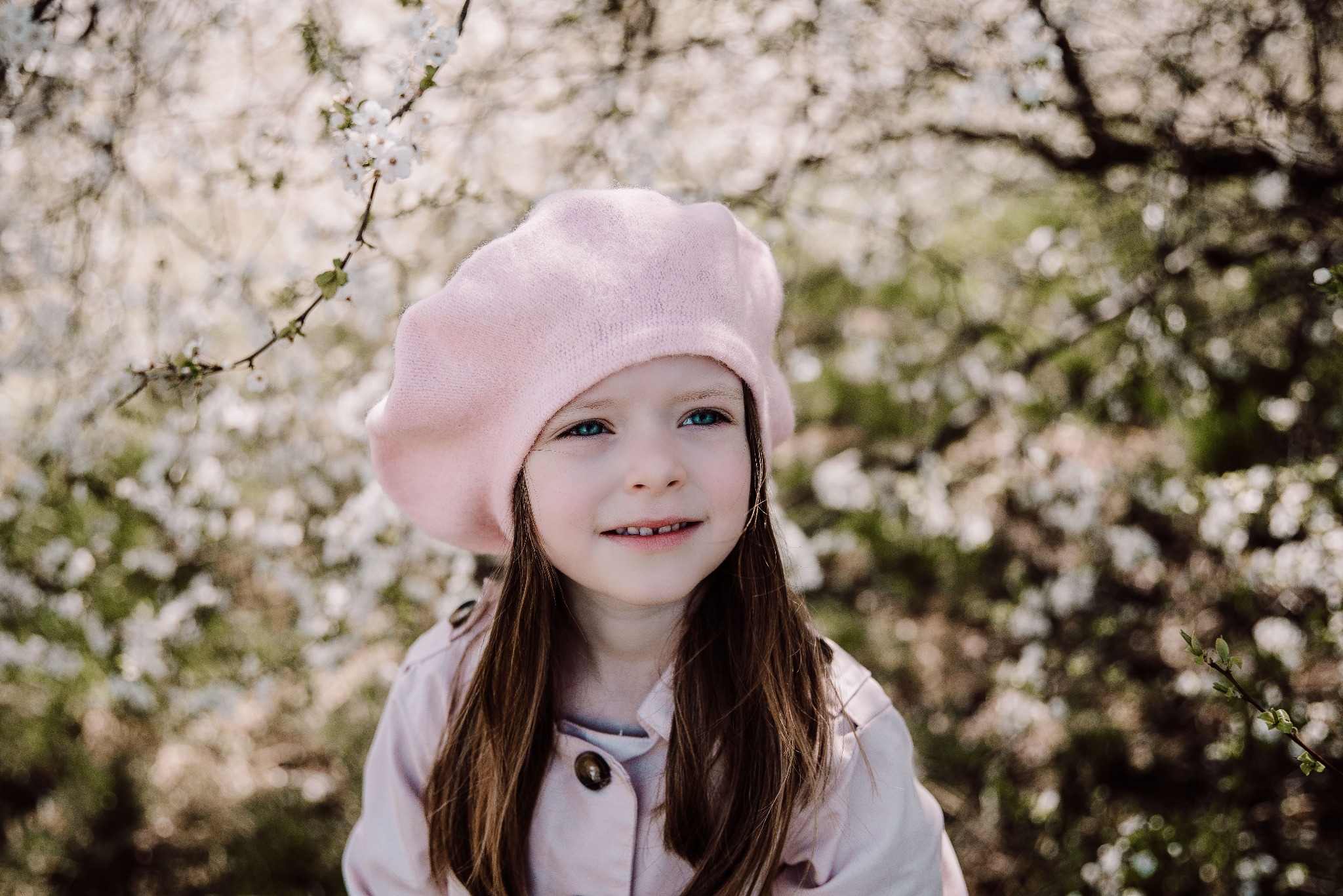 Portret uśmiechniętej dziewczynki w różowym berecie i płaszczu na tle kwitnących gałęzi drzew.