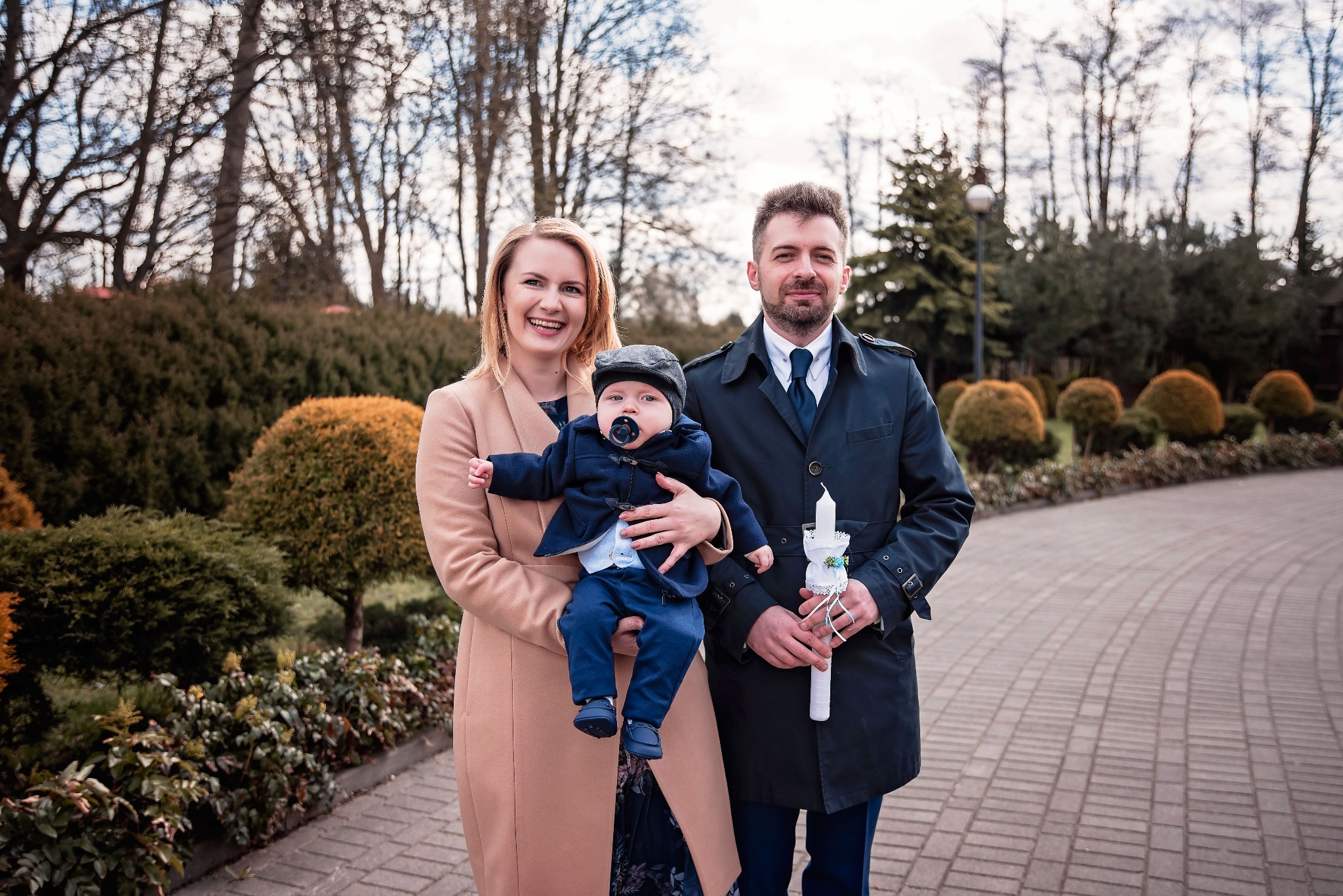 Uśmiechnięta matka trzyma na rękach niemowlę w czapce, ojciec obok trzyma świecę, w tle park z krzewami i brukowana ścieżka.